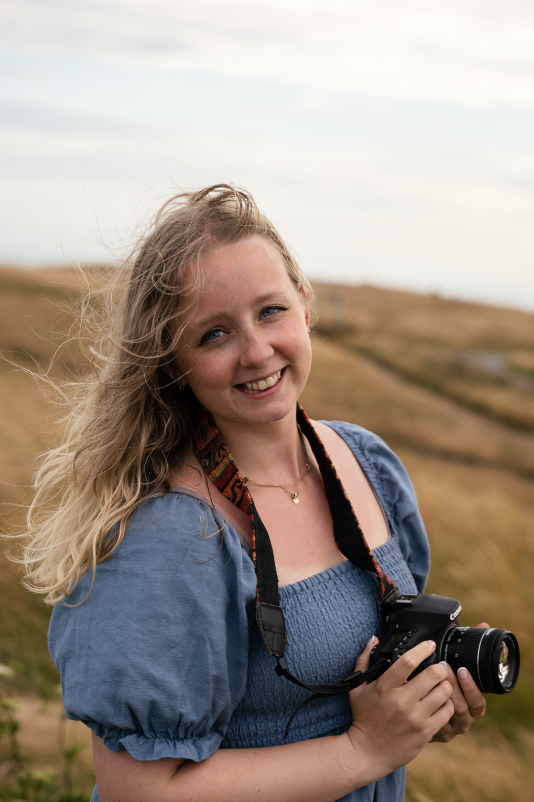 Smiling blonde woman holding camera in golden field on overcast day.