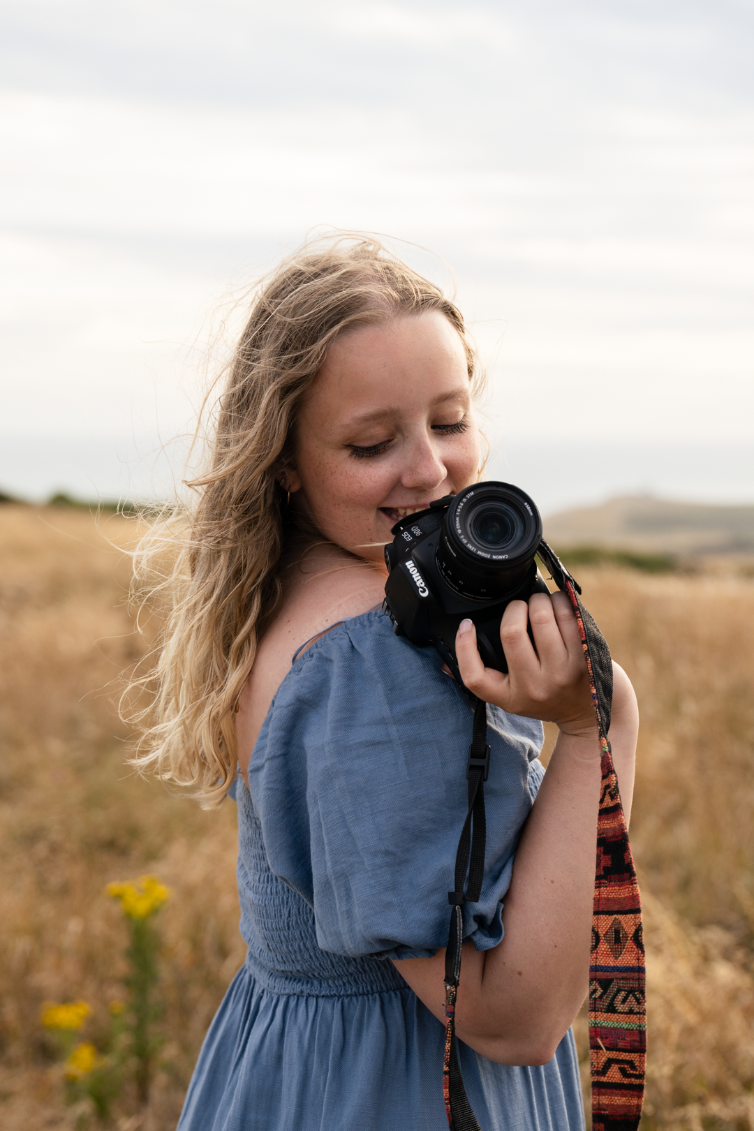 Smiling blonde woman in blue dress holding camera in golden field outdoors.