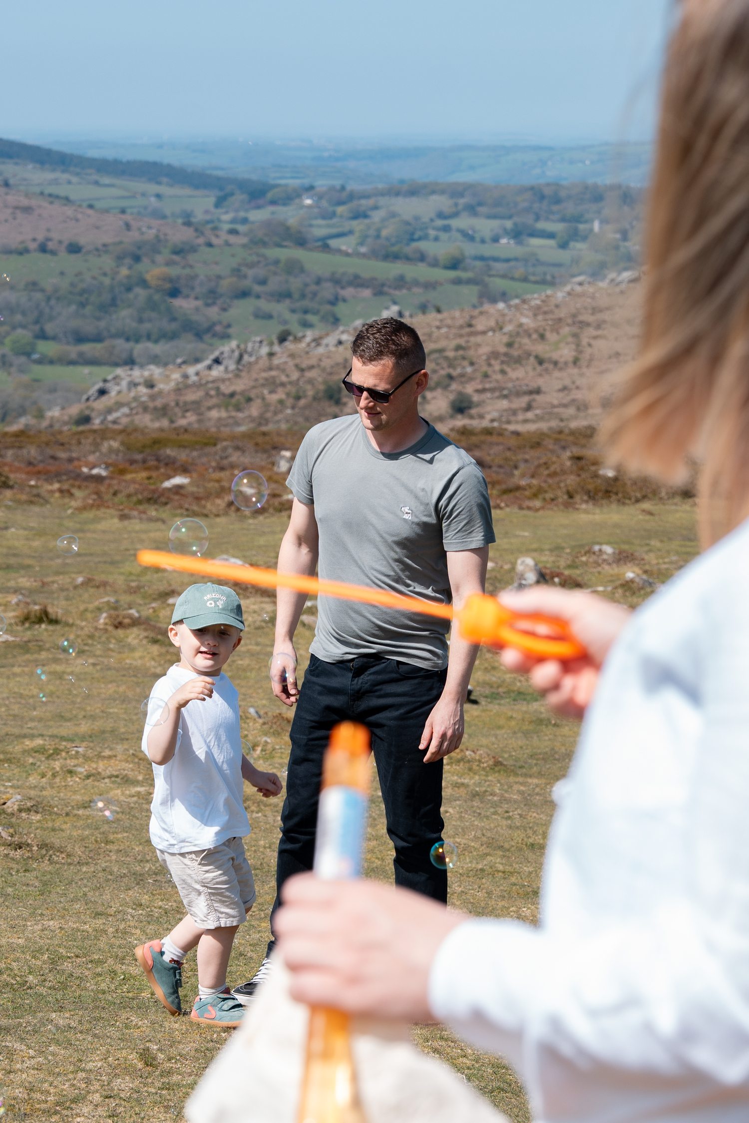 A father and young child play with bubble wands outdoors on a sunny hillside with scenic countryside views.