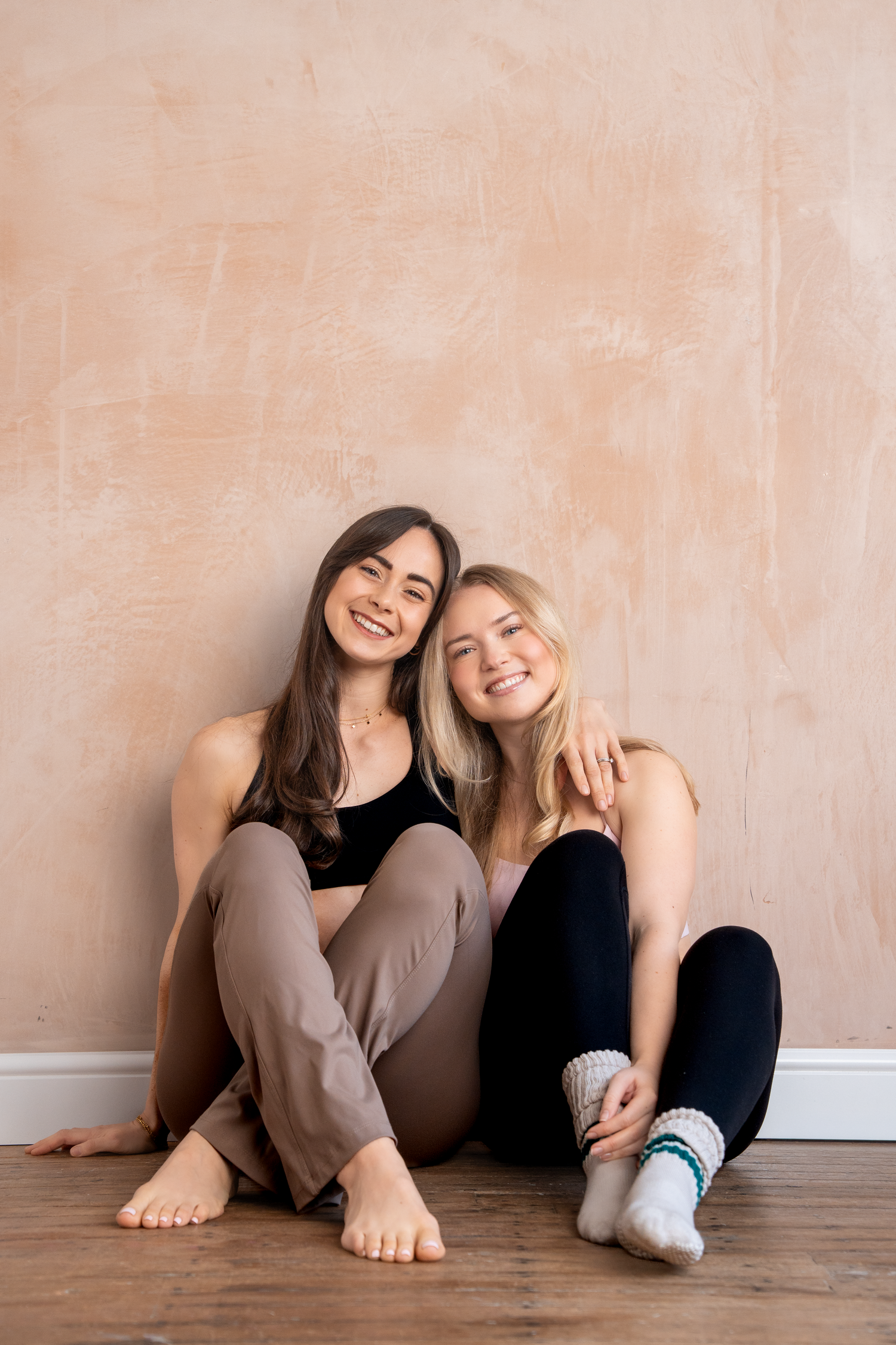 Two smiling young women sitting on wooden floor against beige wall, wearing athletic wear, posing together.