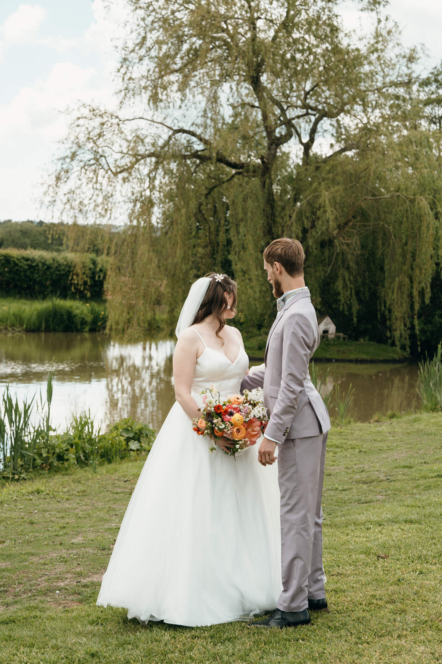 Bride and groom holding hands by a tranquil pond with a willow tree, bride holding colorful bouquet in white gown.