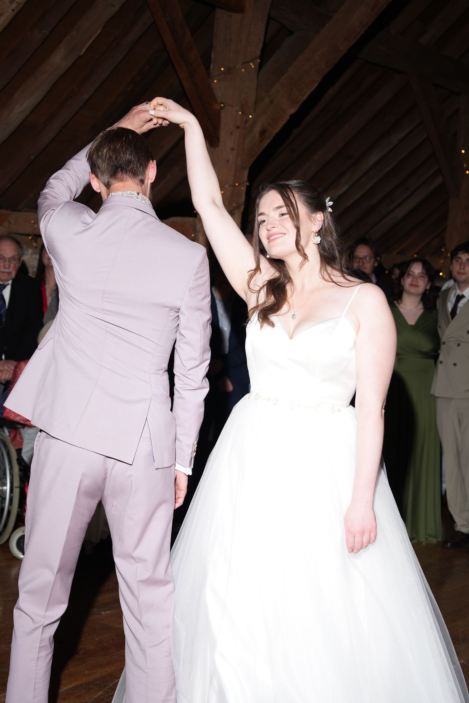Bride in white gown smiles while dancing with groom in pink suit during wedding reception in rustic barn venue.