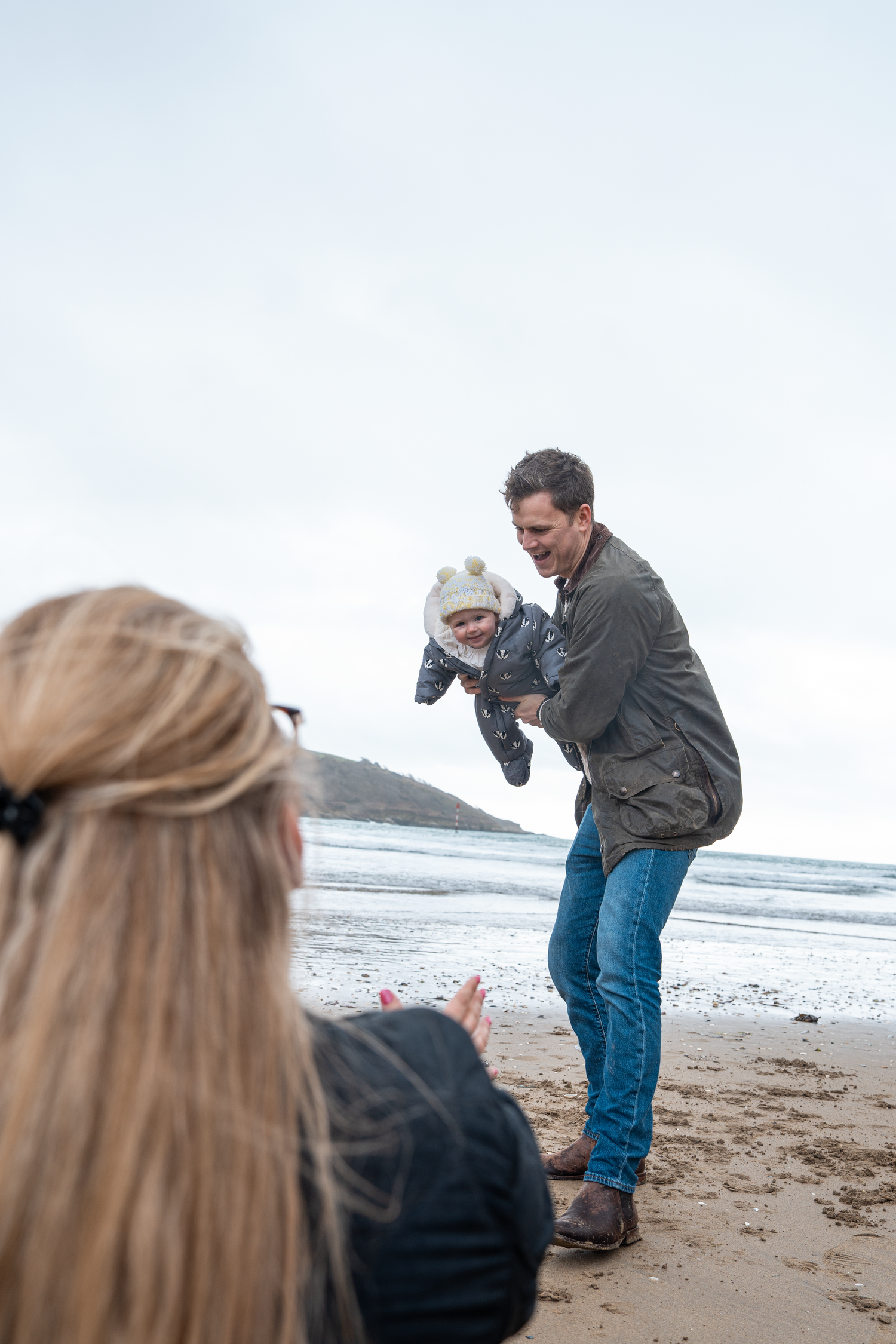 Father playfully lifting baby toward mother on sandy beach.