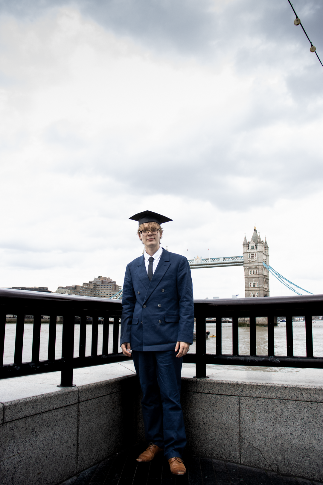 A young man in a navy suit and graduation cap stands before Tower Bridge on a cloudy day.