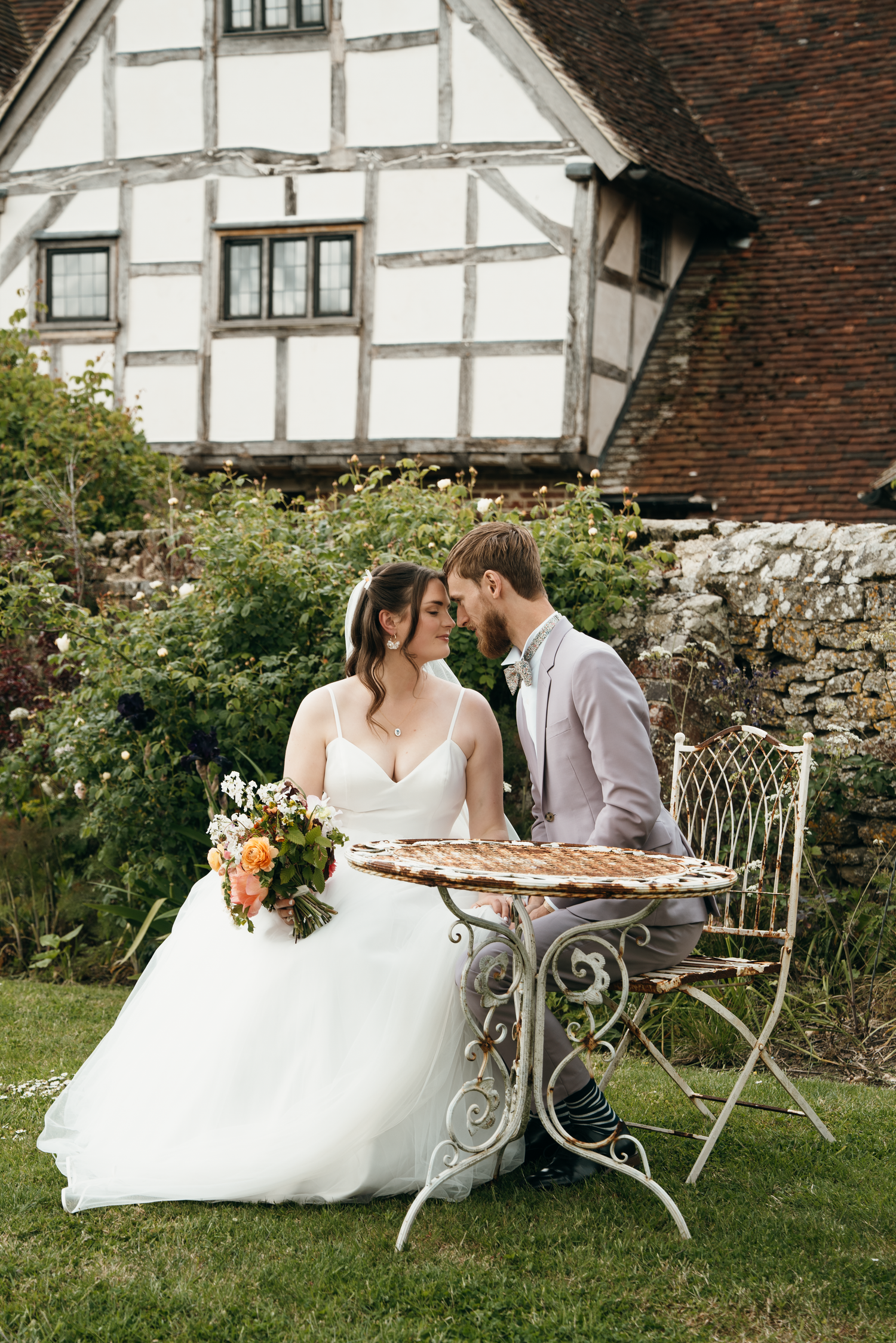Bride and groom share a romantic moment at a vintage iron table in a lush garden beside a Tudor-style country house.