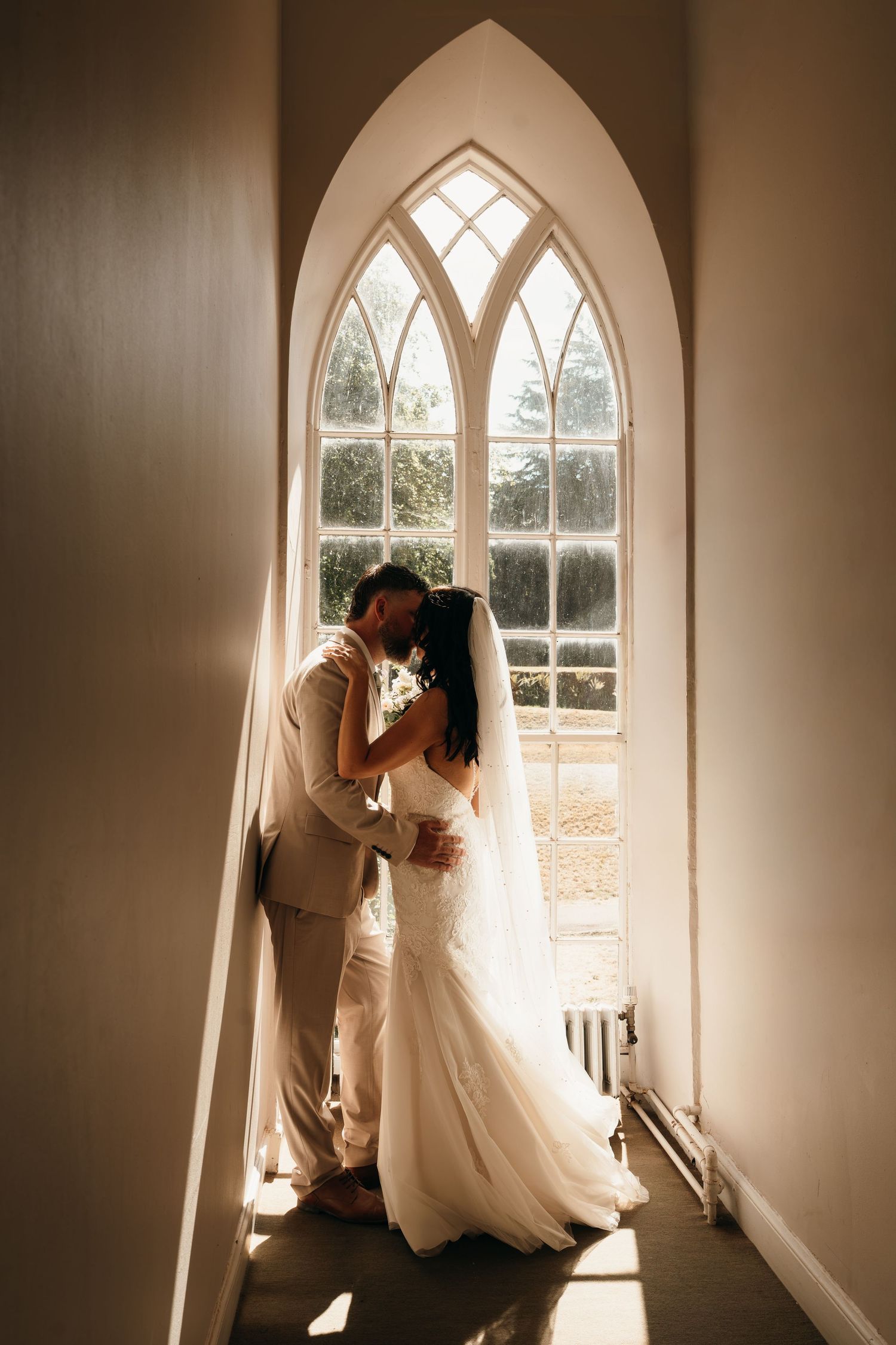 Bride and groom sharing an intimate moment in front of a stunning Gothic arch window, bathed in warm natural light.