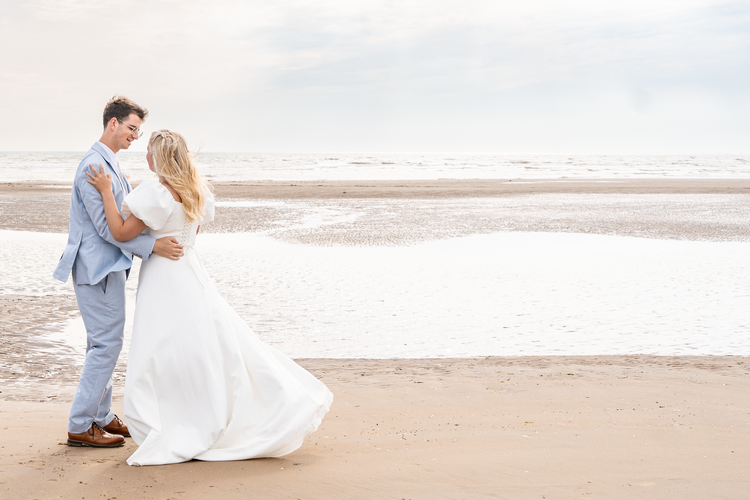 Bride and groom embracing on sandy beach, white dress flowing in the breeze by the ocean.