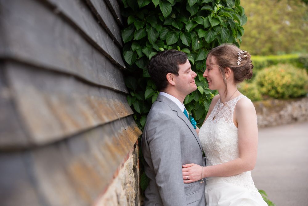 Bride and groom share a romantic moment leaning against a stone wall covered in ivy at their wedding.