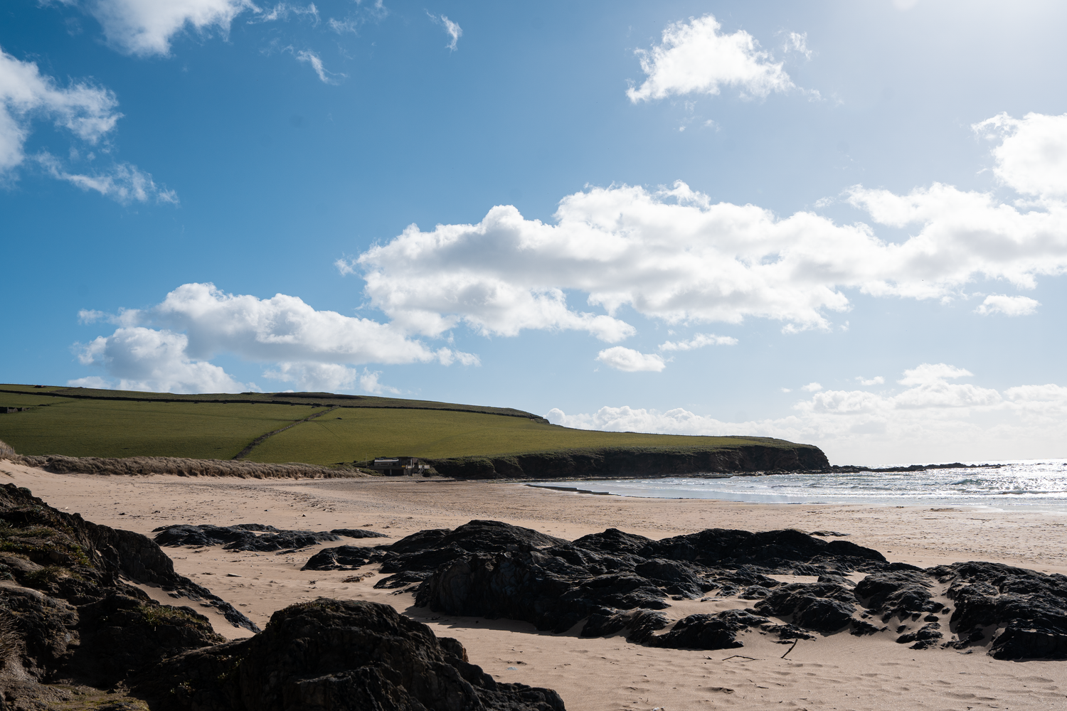 Sandy beach with rocky foreground, green hillside, and blue sky with white clouds on a sunny day.