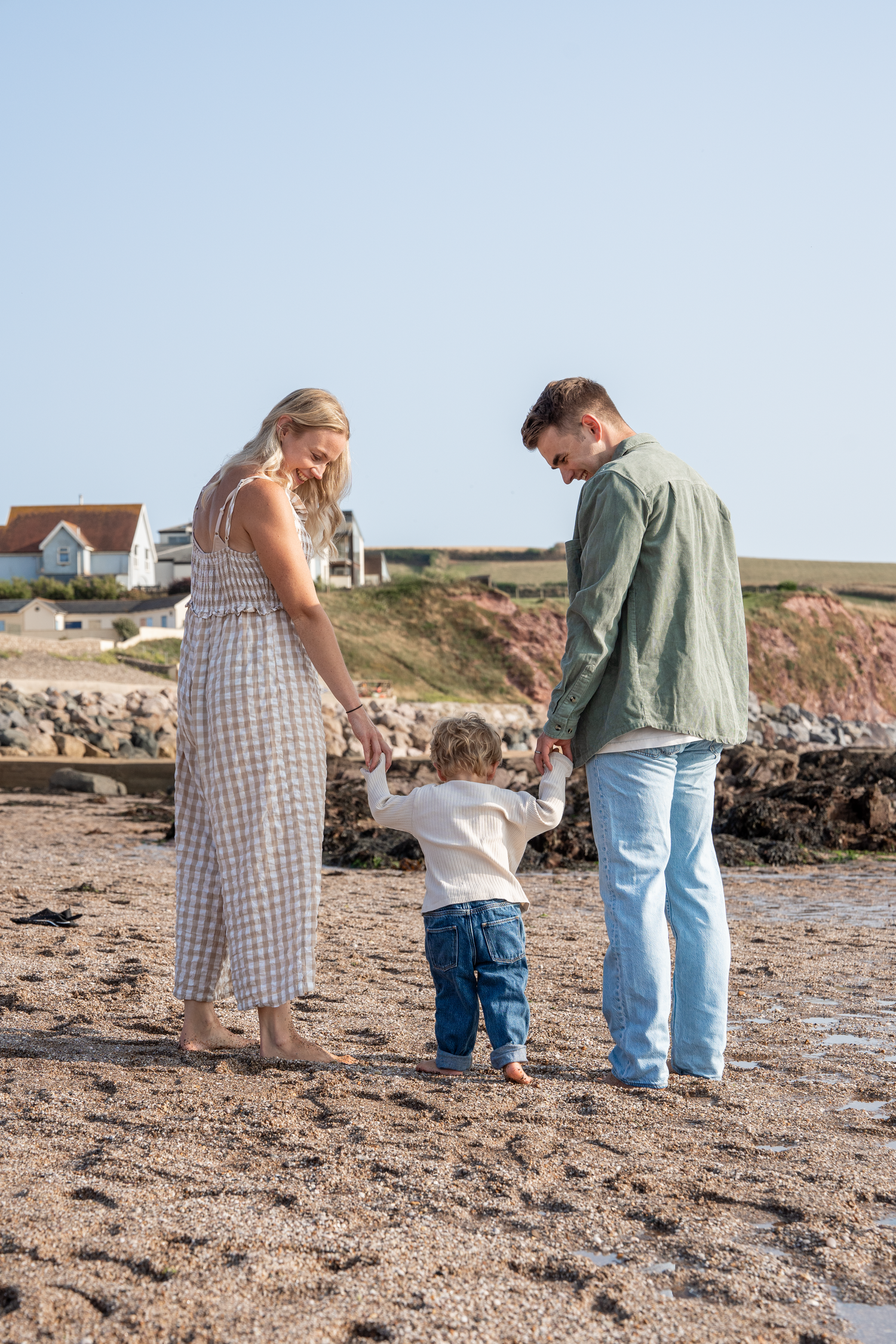 Young family with toddler holding parents hands on a sunny pebble beach with coastal houses behind.