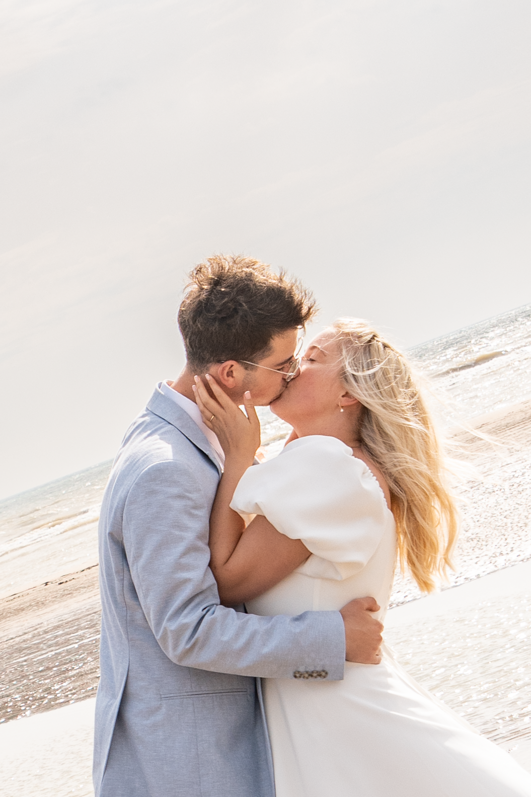A bride and groom share a romantic kiss on a sunlit beach, with the ocean shimmering in the background.