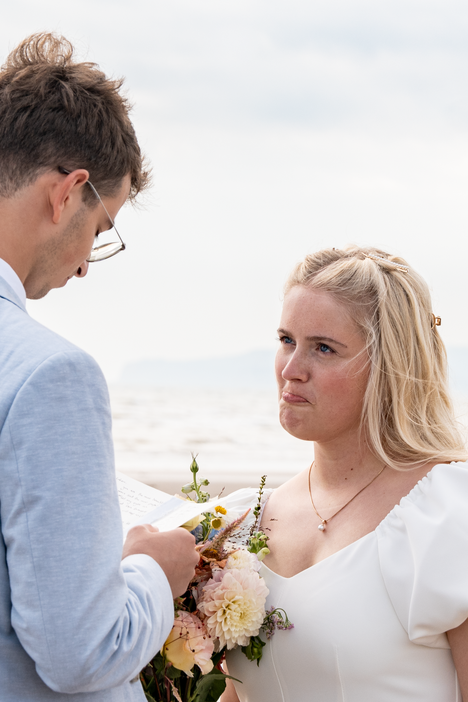 Emotional bride holding floral bouquet listens to groom reading vows at outdoor beach wedding ceremony.