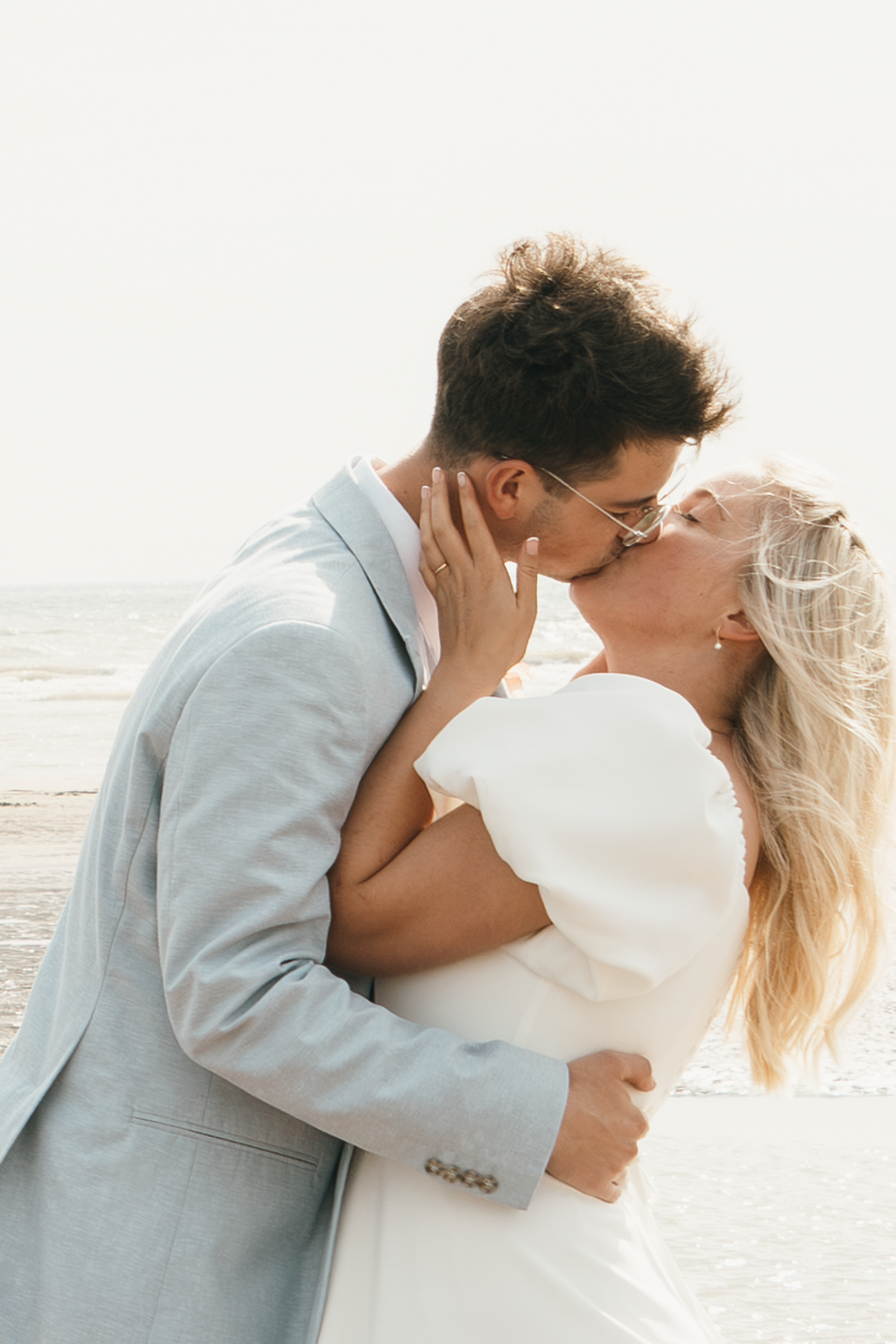 A couple shares a romantic kiss on the beach, with the groom in a light blue suit and bride in a white dress.