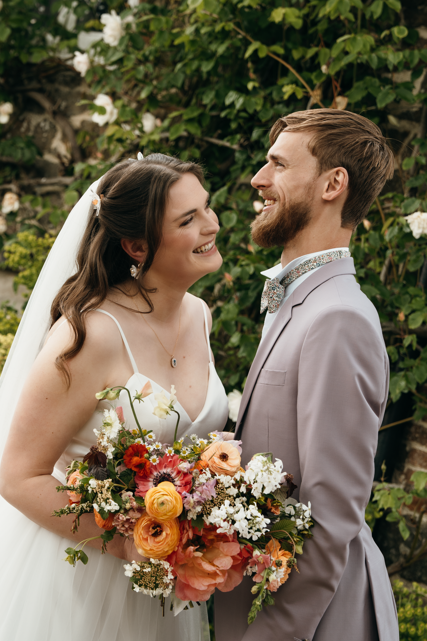 A smiling bride and groom pose outdoors with a colorful bouquet of ranunculus and wildflowers on their wedding day.