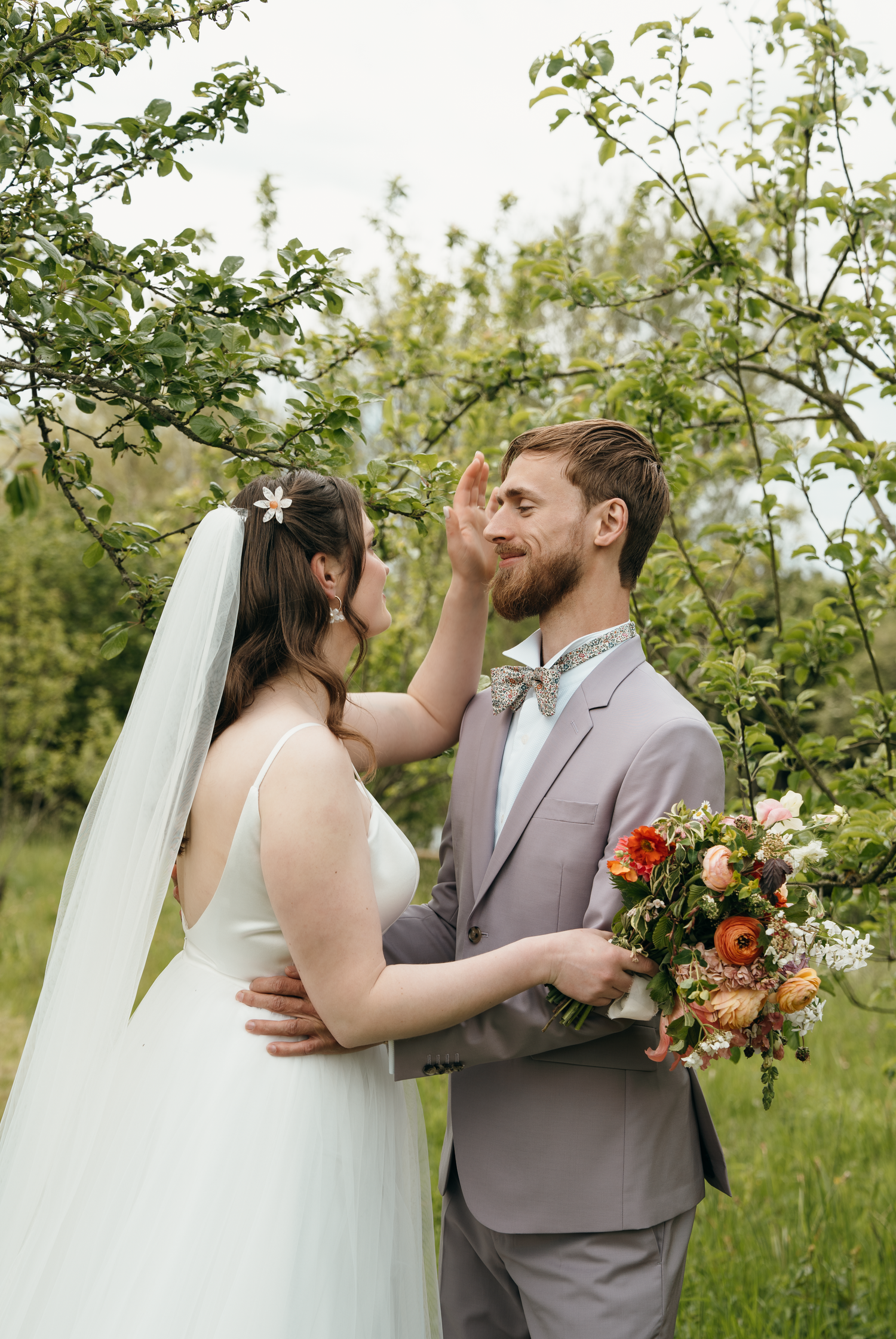 A bride and groom share a tender moment outdoors under green trees, groom holding a colorful bouquet in a gray suit.