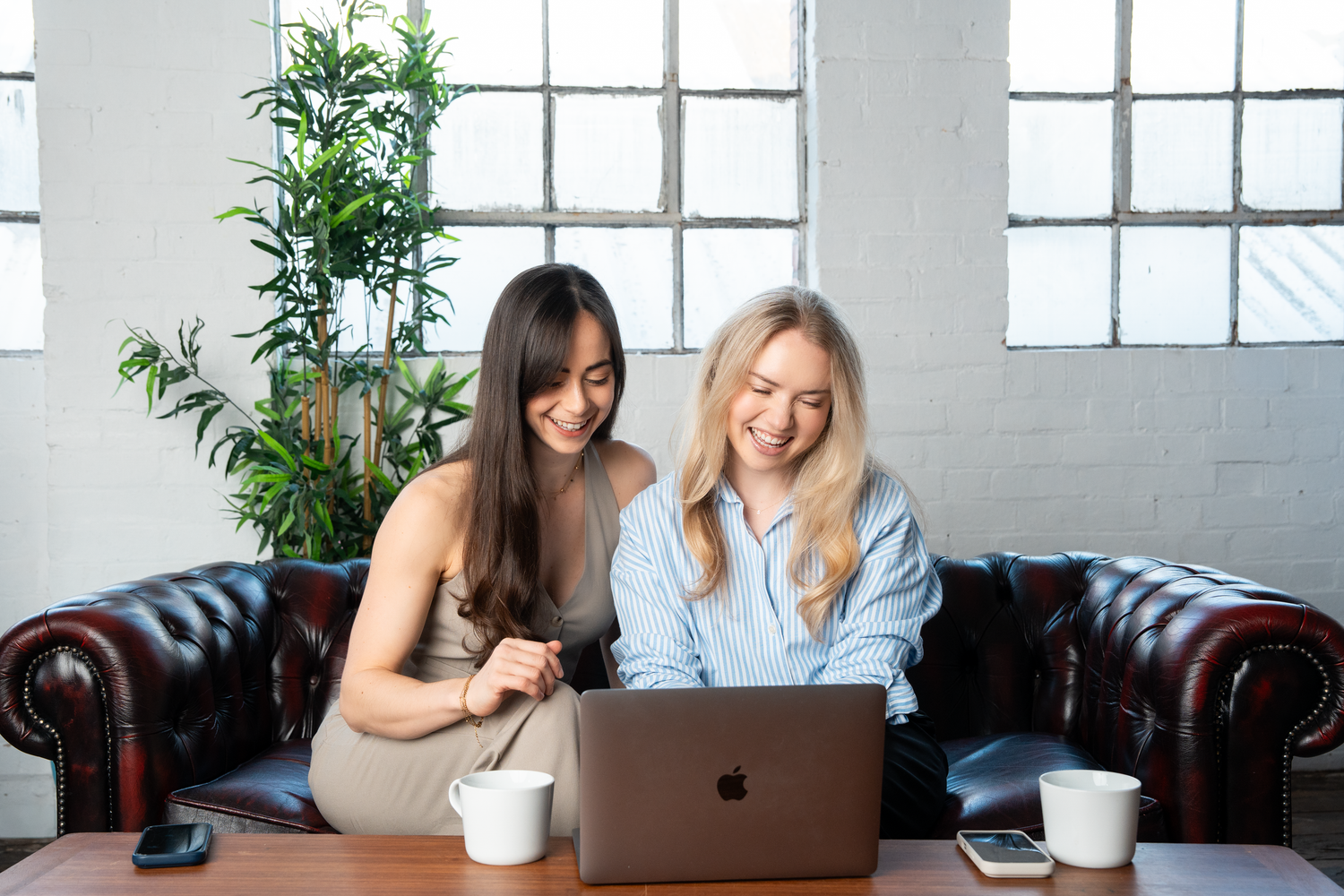 Two smiling women collaborating on a MacBook laptop while sitting on a leather sofa in a bright modern office.