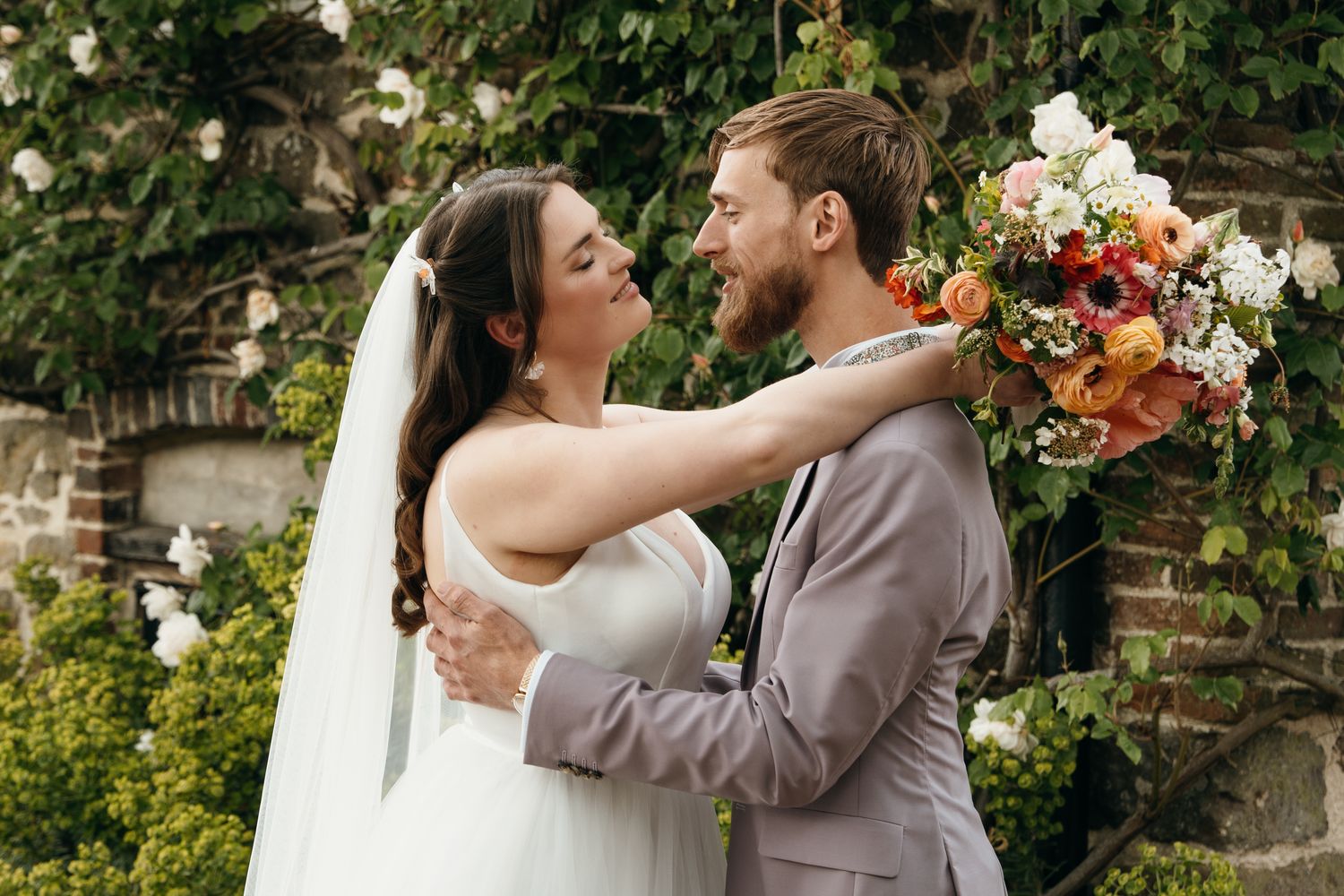 Bride and groom share a romantic moment, she holds a colorful bouquet while embracing outdoors near lush greenery.