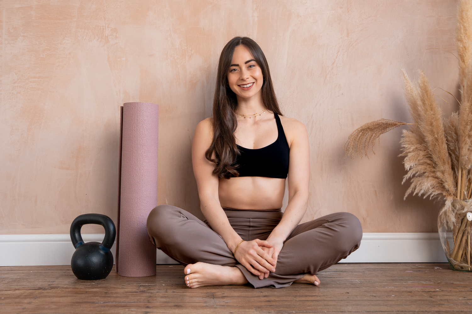 Smiling woman in black sports bra sitting cross-legged on floor near yoga mat and kettlebell.