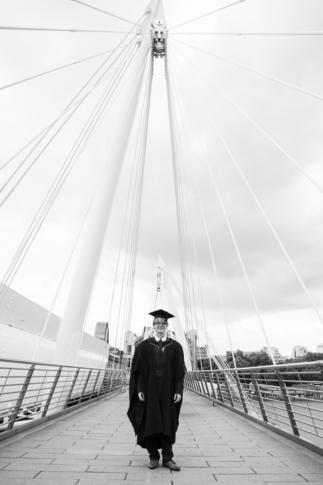 A graduate in cap and gown stands confidently on a modern suspension bridge in a black and white photo.
