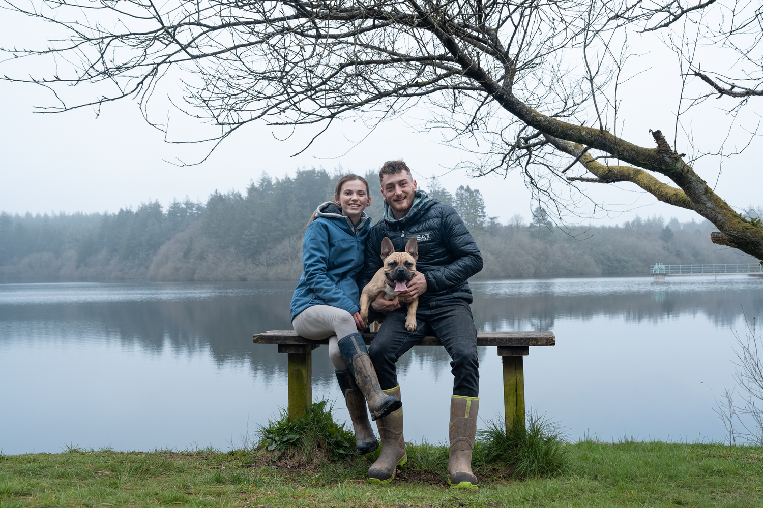 Couple sitting on a bench by a misty lake holding a small dog, wearing rain jackets and wellies outdoors.
