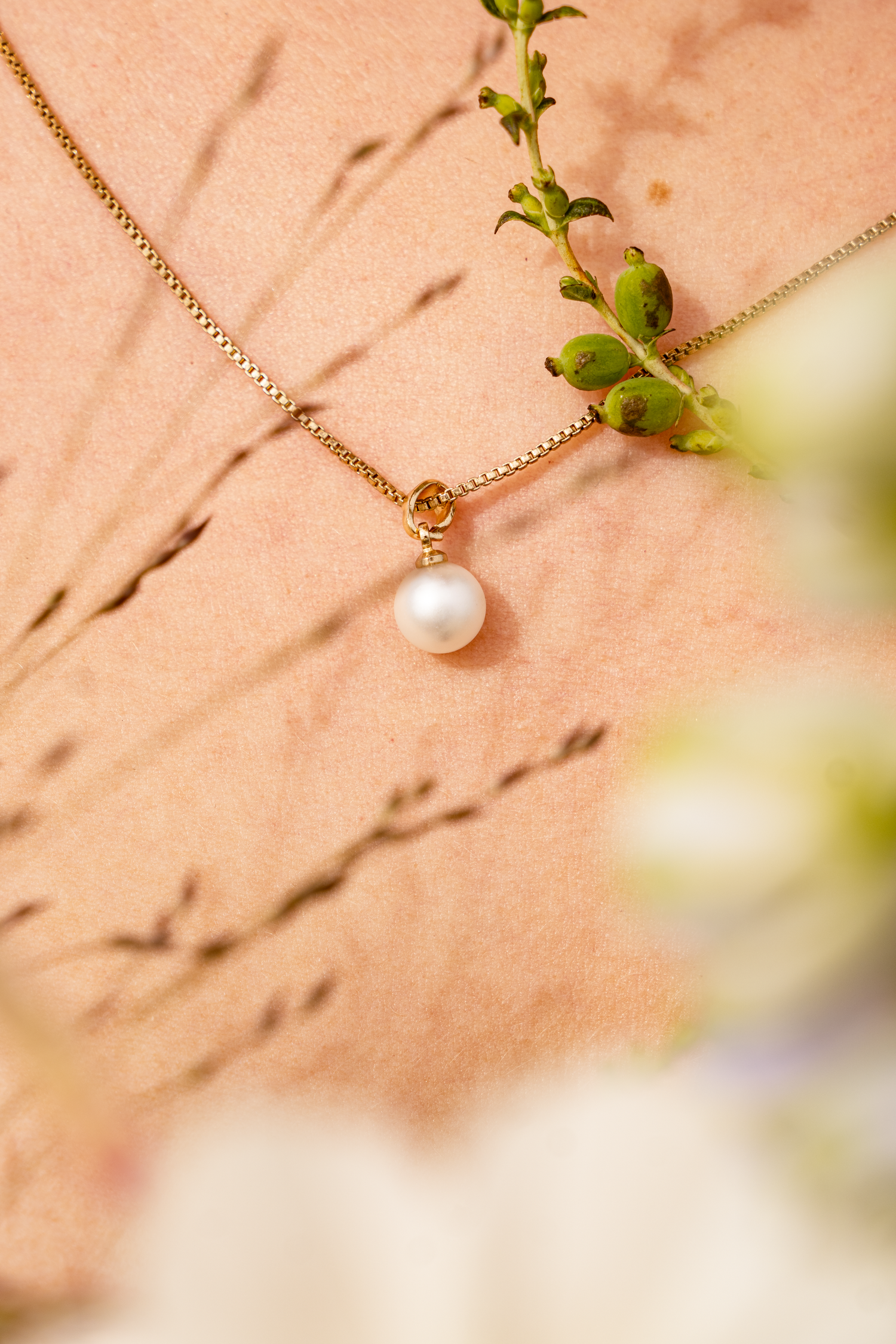 Elegant gold pearl pendant necklace on chain displayed with green botanical stems.