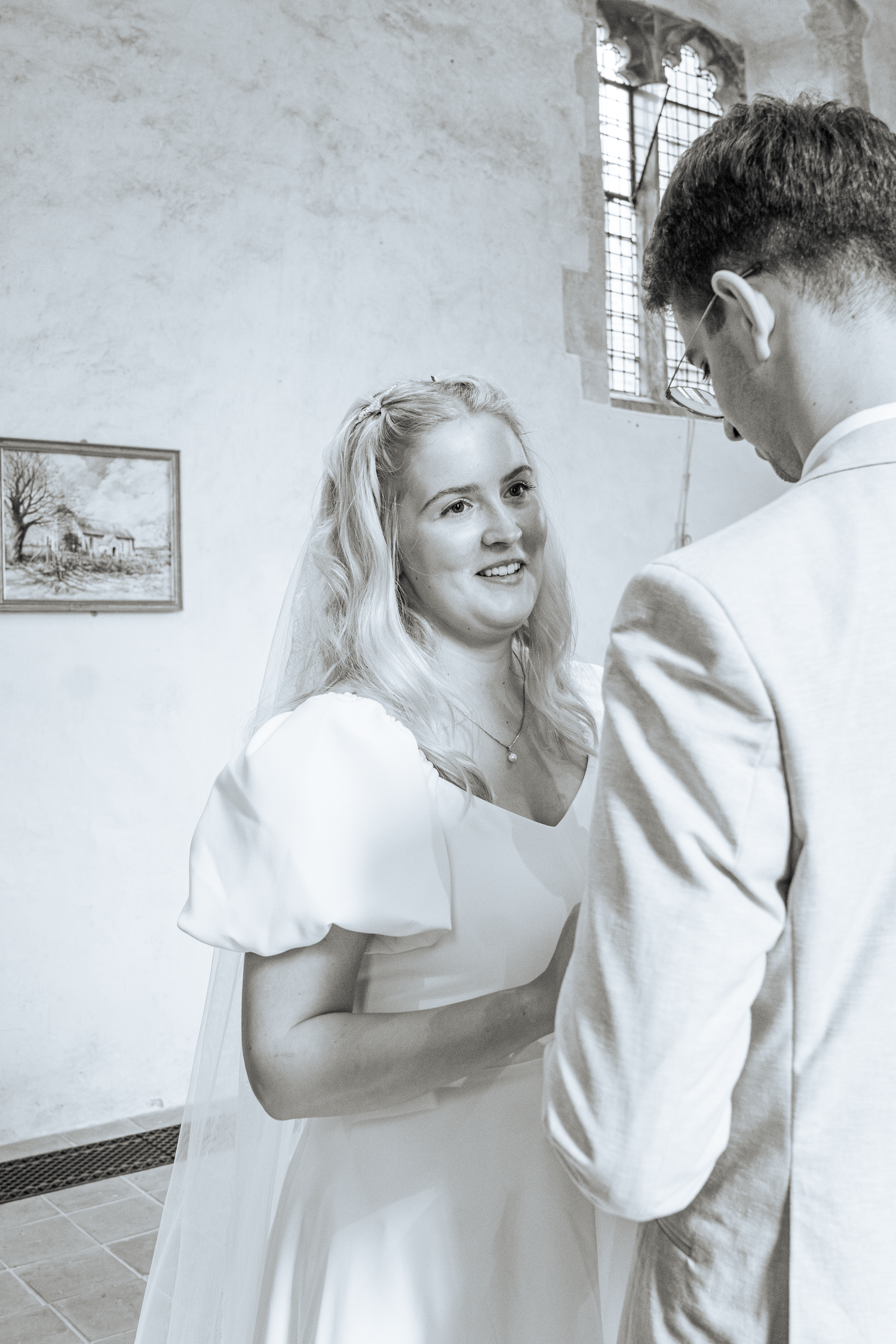 Black and white photo of smiling blonde bride in veil looking up at groom inside historic church.