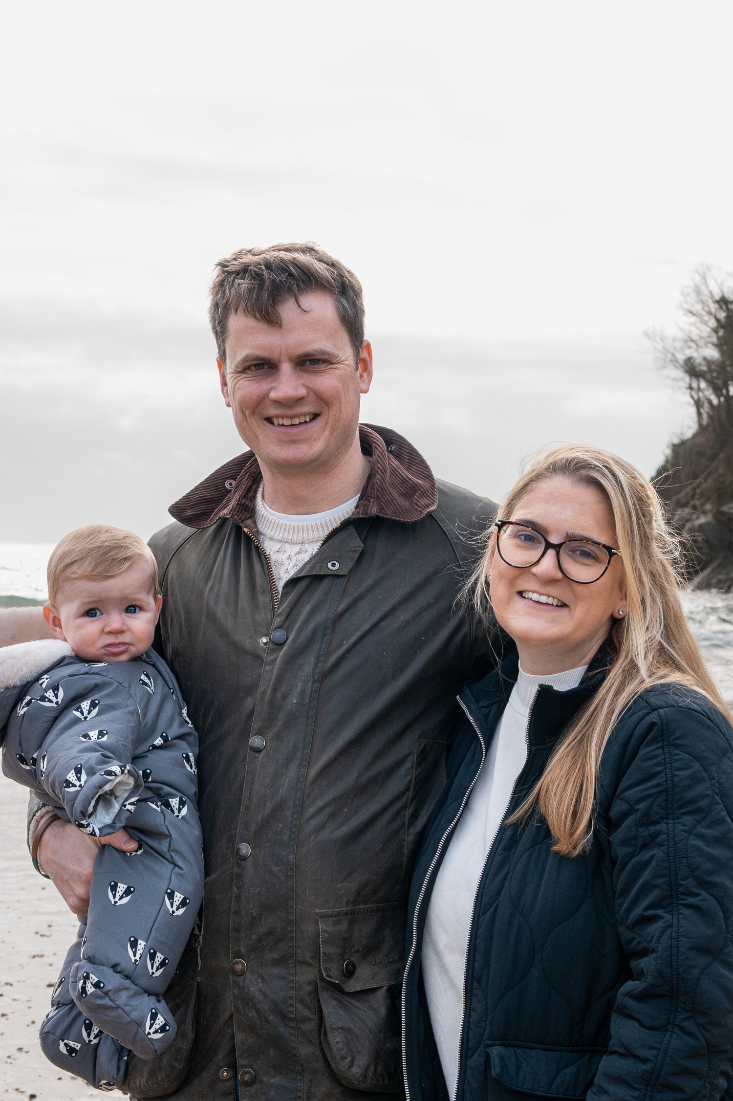 Smiling family portrait with father holding baby in badger-print suit and mother with glasses on beach.