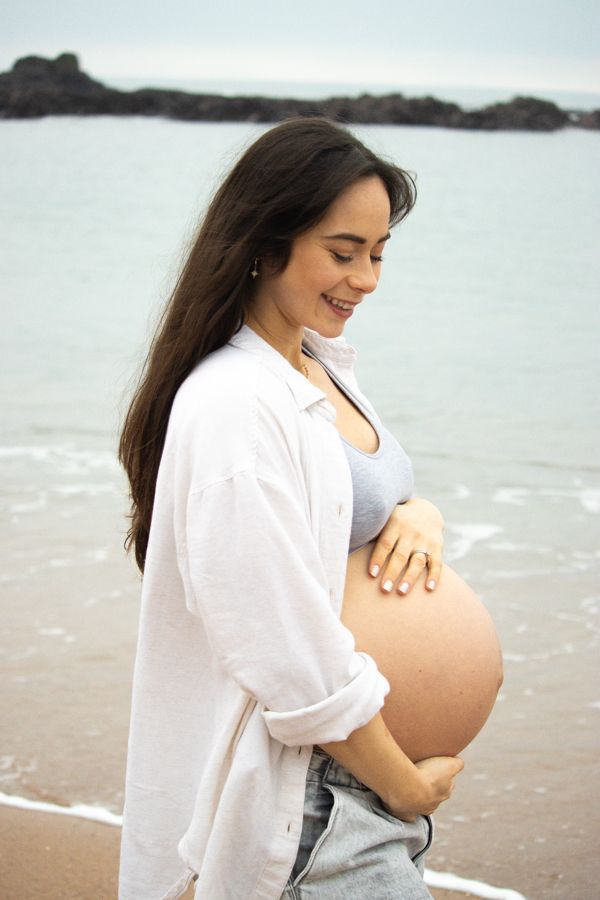Smiling pregnant woman in white shirt tenderly holding her bare baby bump on a beach.