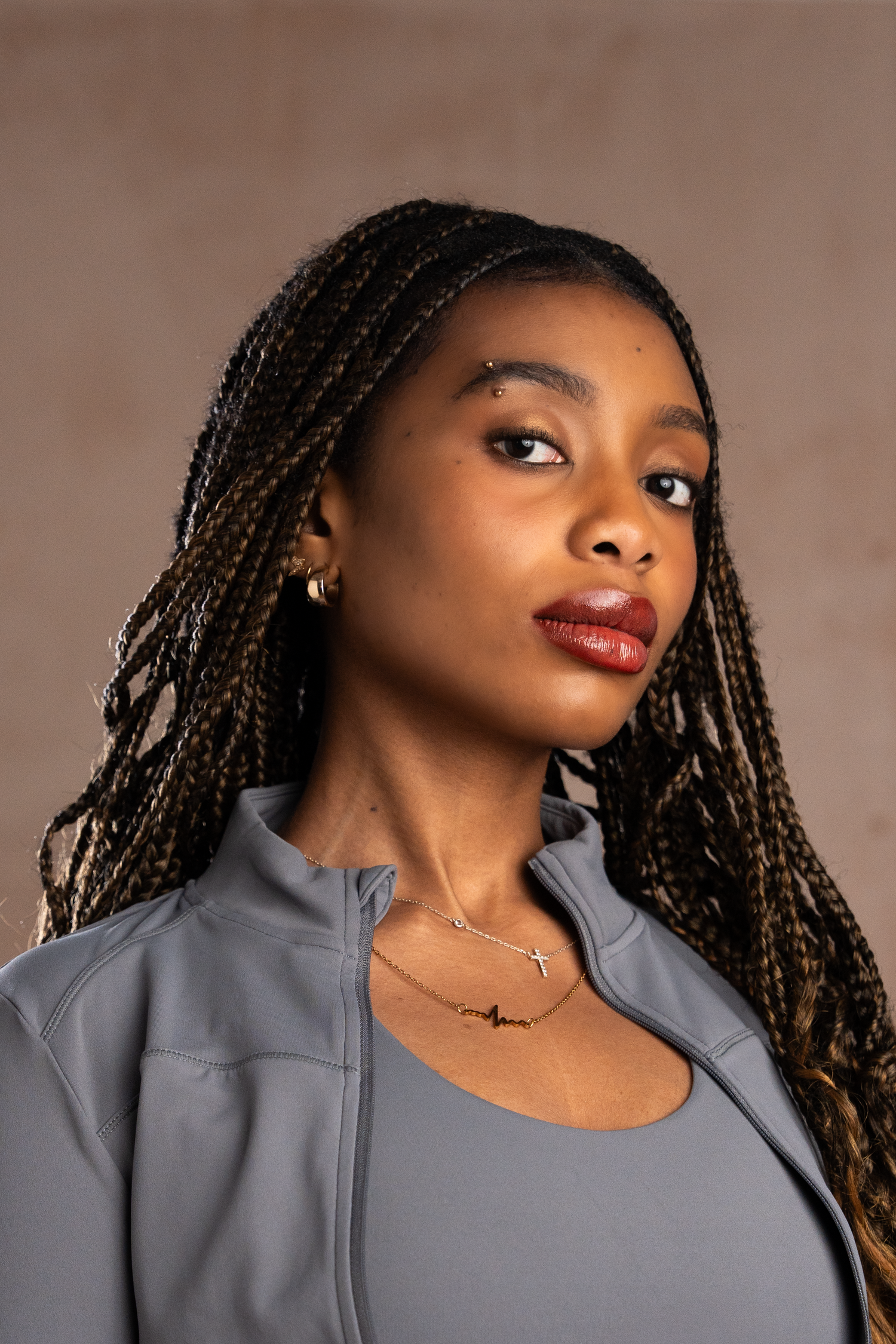 Young woman with braids wearing a grey zip-up jacket and gold cross necklace against a lime wash wall.