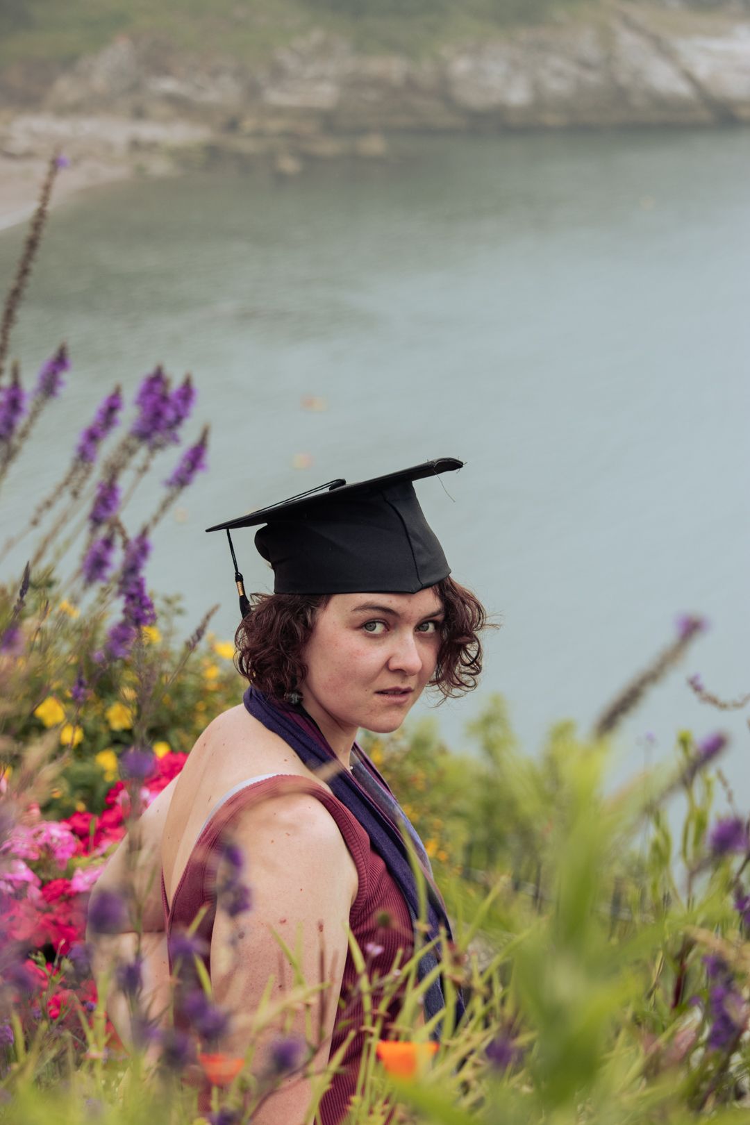 Graduate wearing black graduation cap poses among vibrant purple and pink flowers with coastal backdrop.