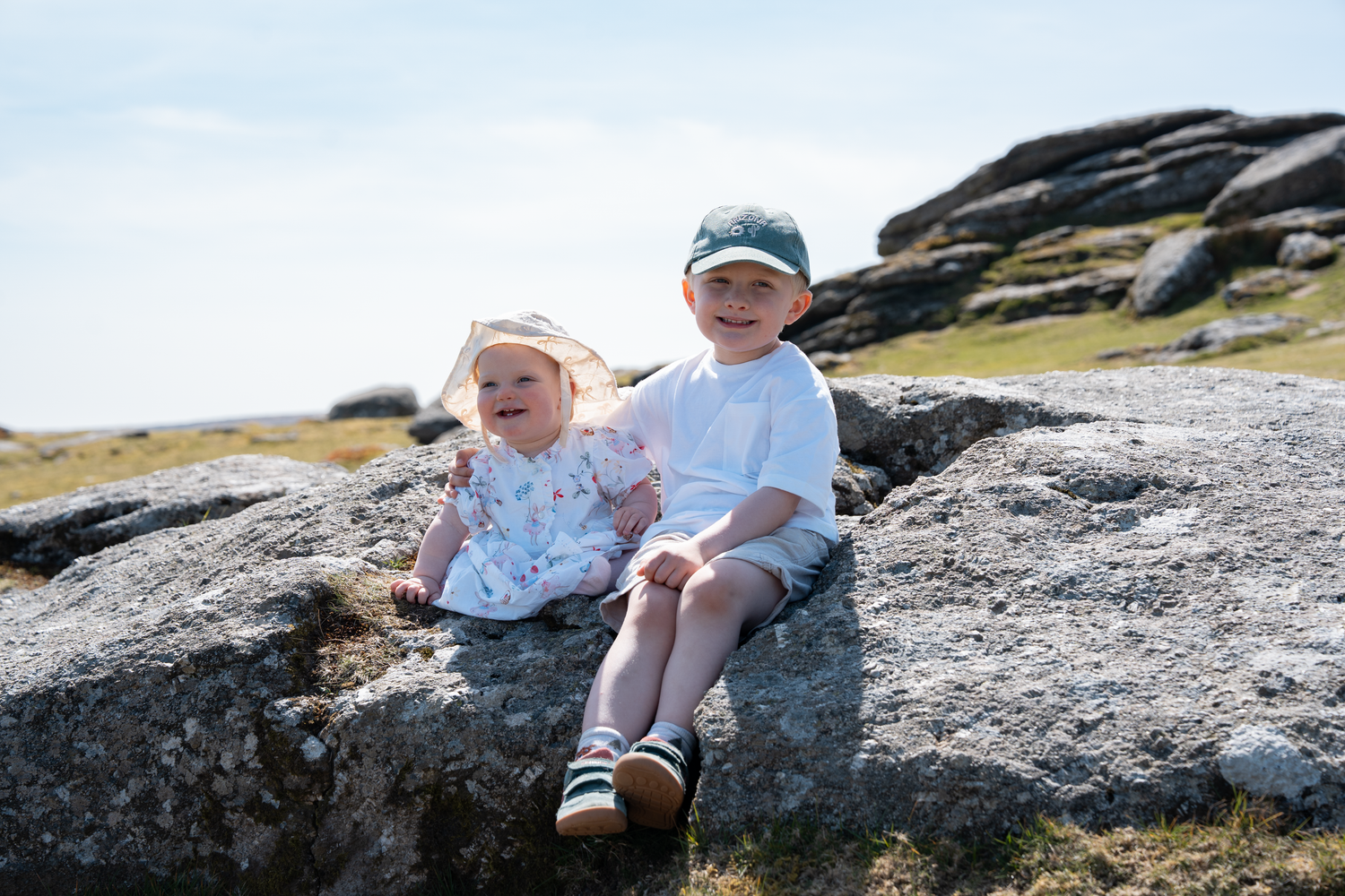 Two young children sitting on a large granite rock outdoors, wearing summer hats and white clothing on a sunny day.