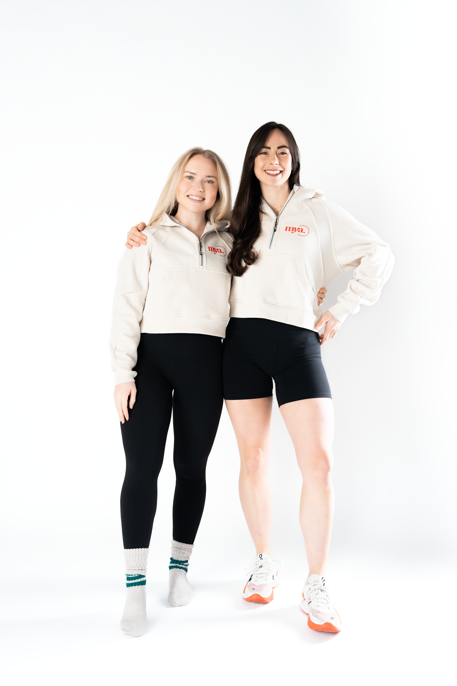 Two women in matching cream hoodies and black shorts smile together against a white background.