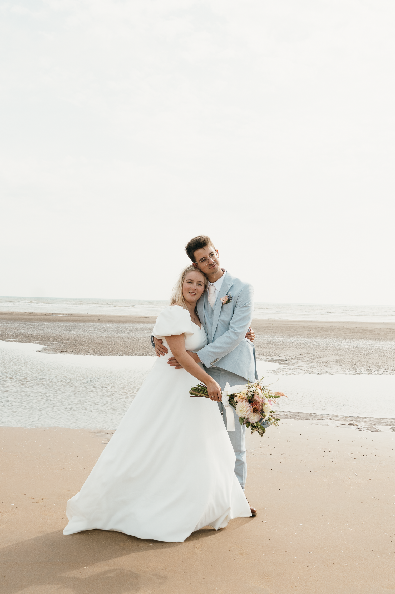 A smiling bride in a white gown and groom in a light blue suit embrace on a sandy beach holding a floral bouquet.