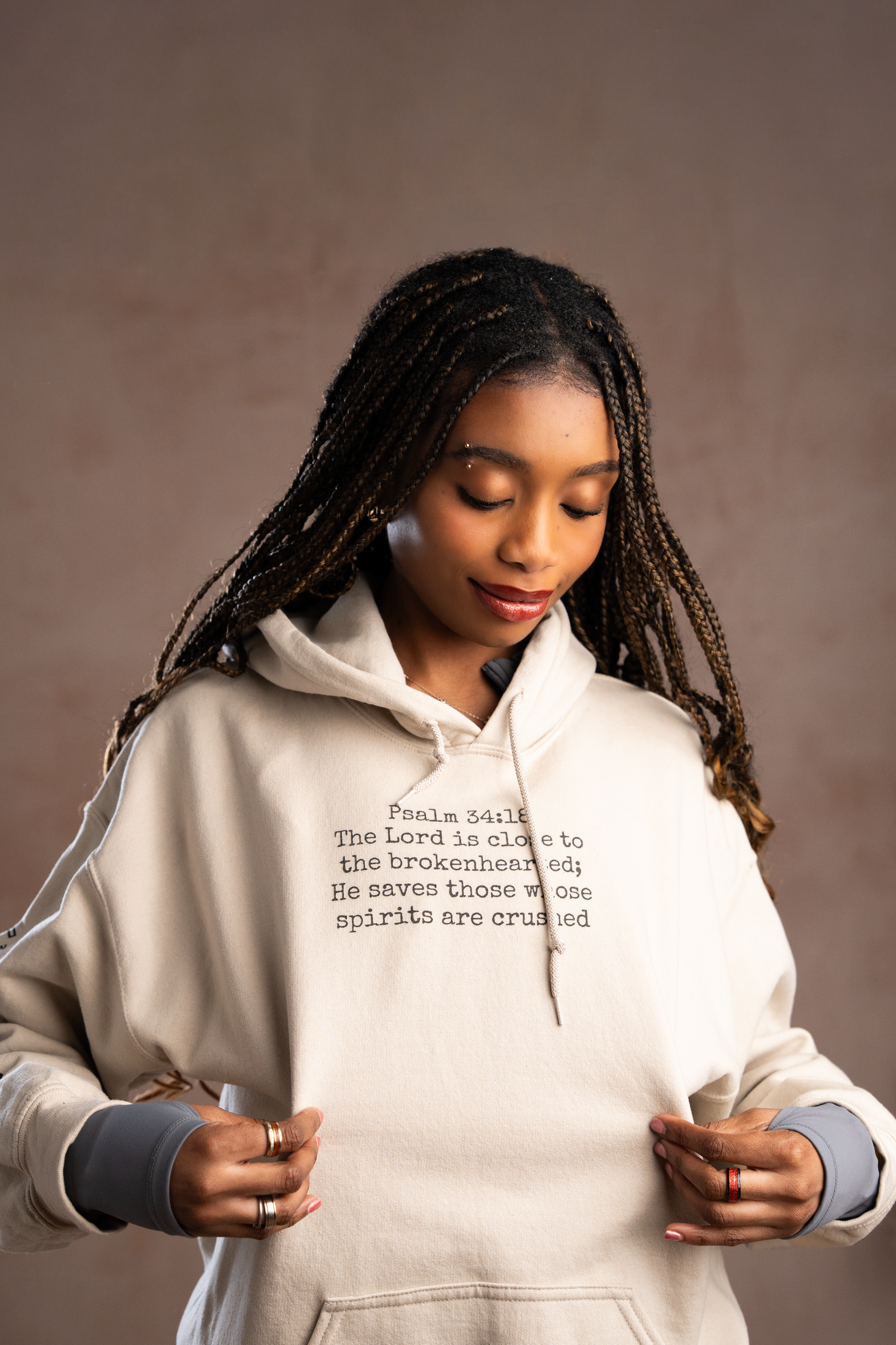 Young woman wearing a cream hoodie with Psalm 34:18 Bible verse, featuring braids and gold rings.