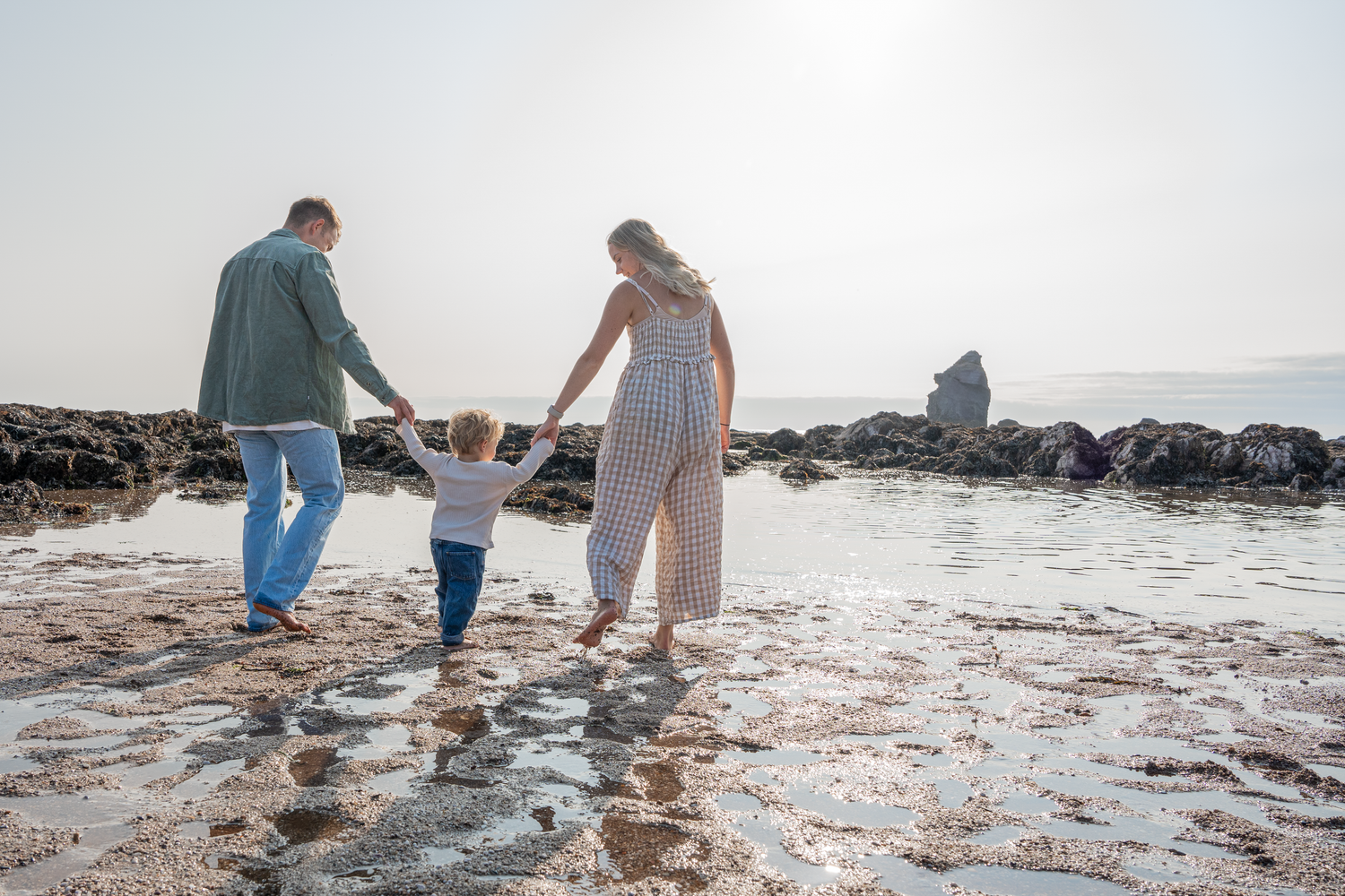 Young family of three holding hands walking on rocky beach shoreline at sunset.