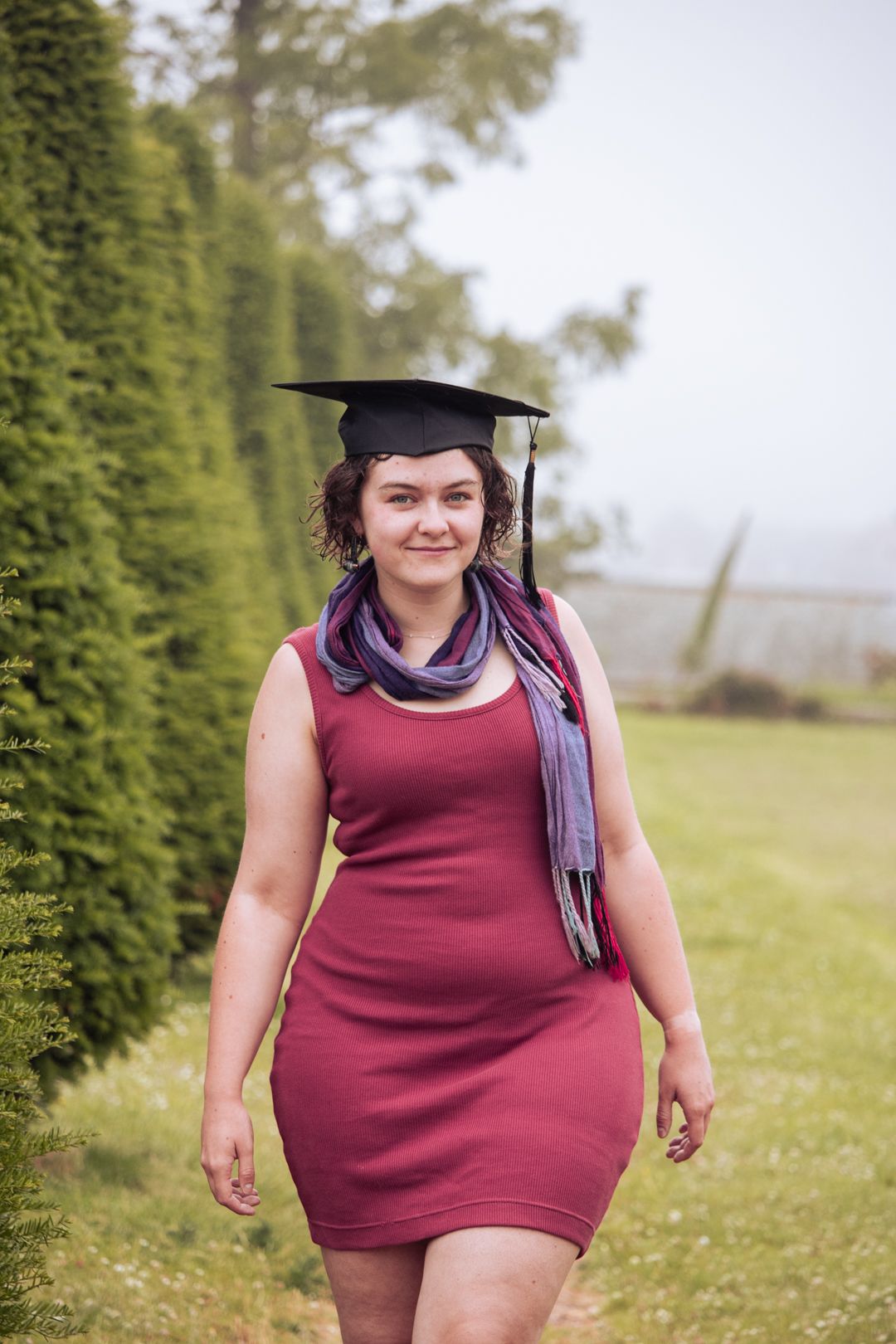 Smiling graduate in burgundy dress and graduation cap walks along misty garden path between tall hedges.