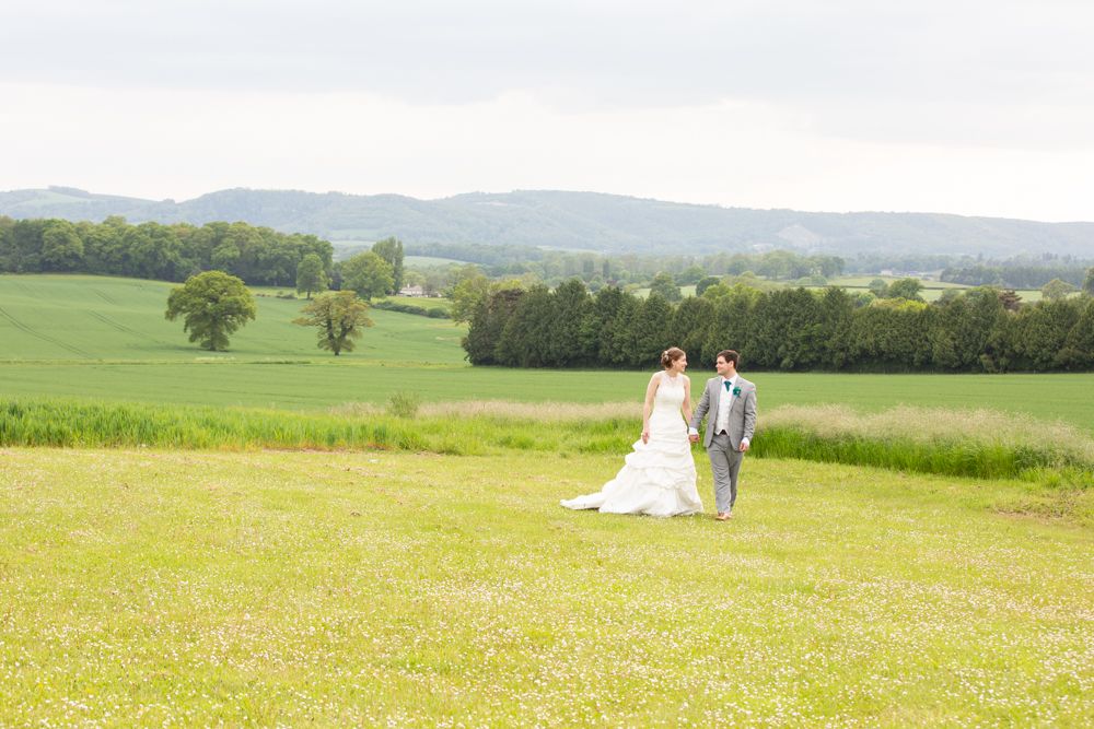 Bride and groom walking through a lush green meadow with rolling hills backdrop.
