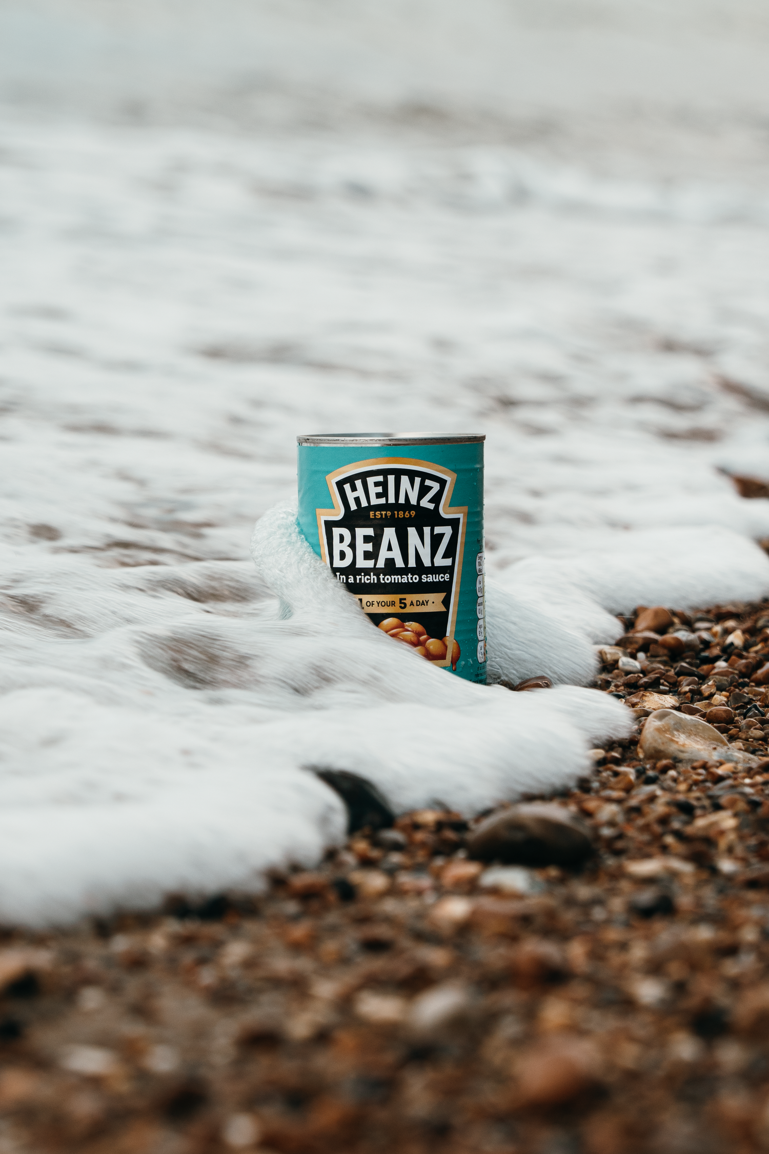 A Heinz Beanz can sitting on a pebble beach surrounded by white sea foam waves.