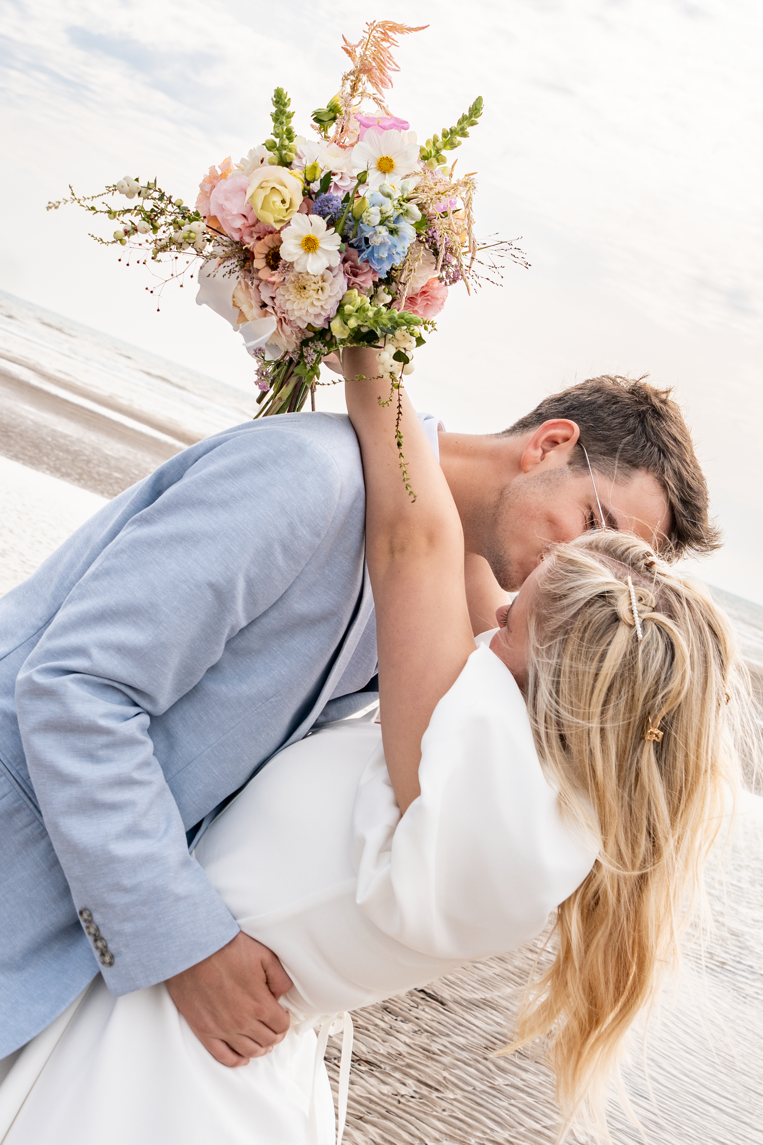 Bride and groom share a romantic kiss on the beach, with a colorful spring bouquet held above them.