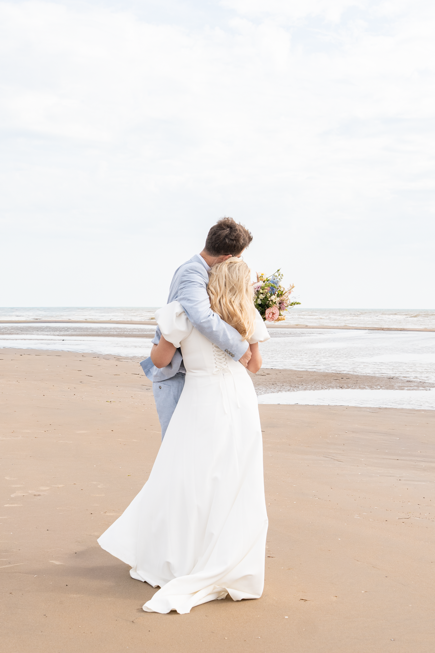 Bride and groom embracing on a sandy beach, bride holding a bouquet in a white gown, groom in a light blue suit.