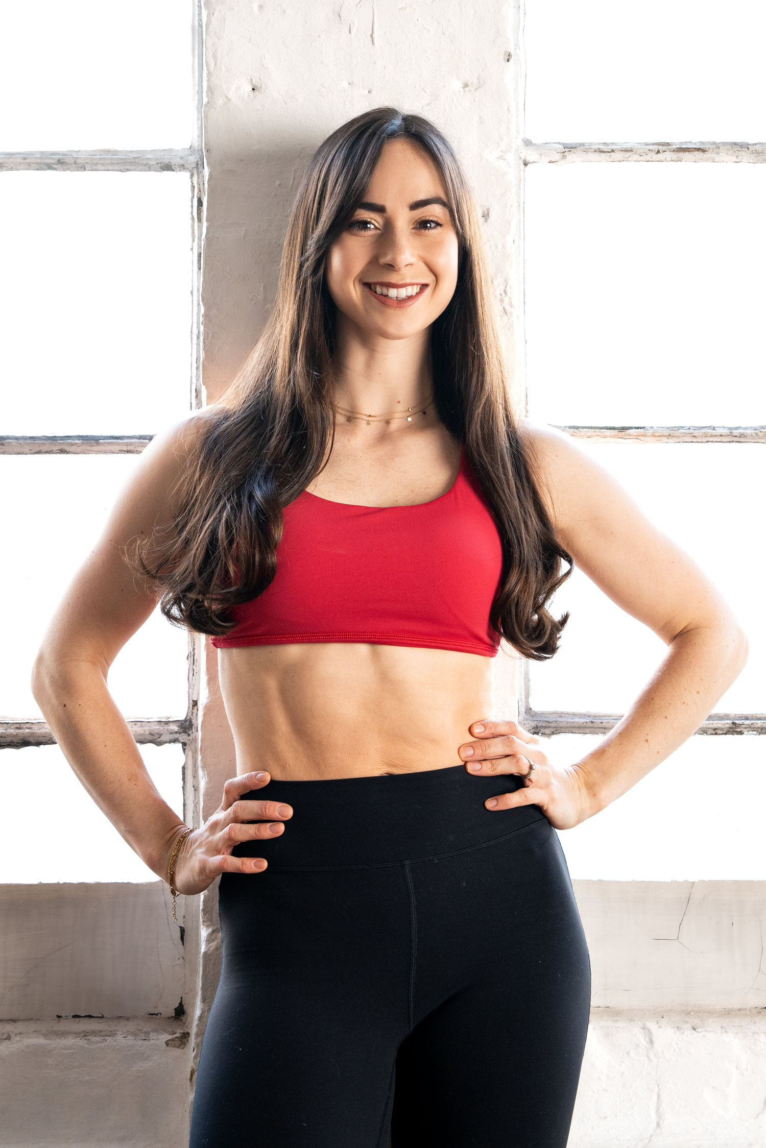 Fit brunette woman in red sports bra and black leggings showing toned abs by window.