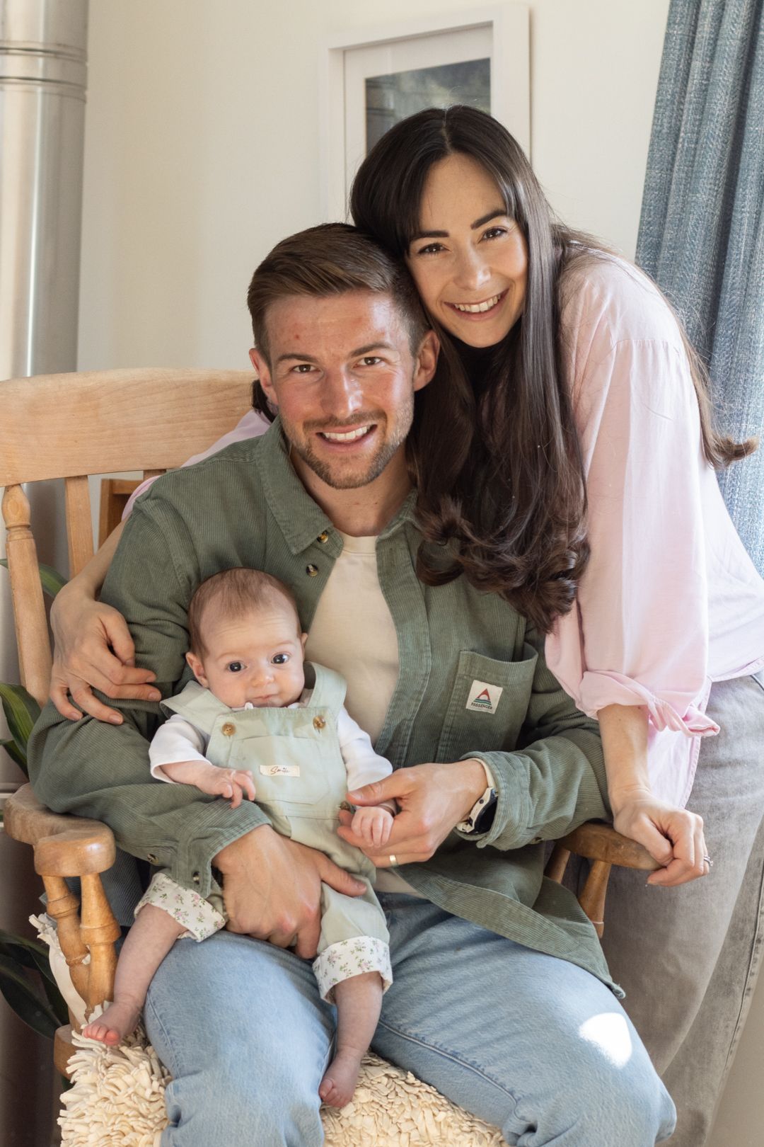 Smiling family portrait with father holding newborn in rocking chair, mother leaning in.