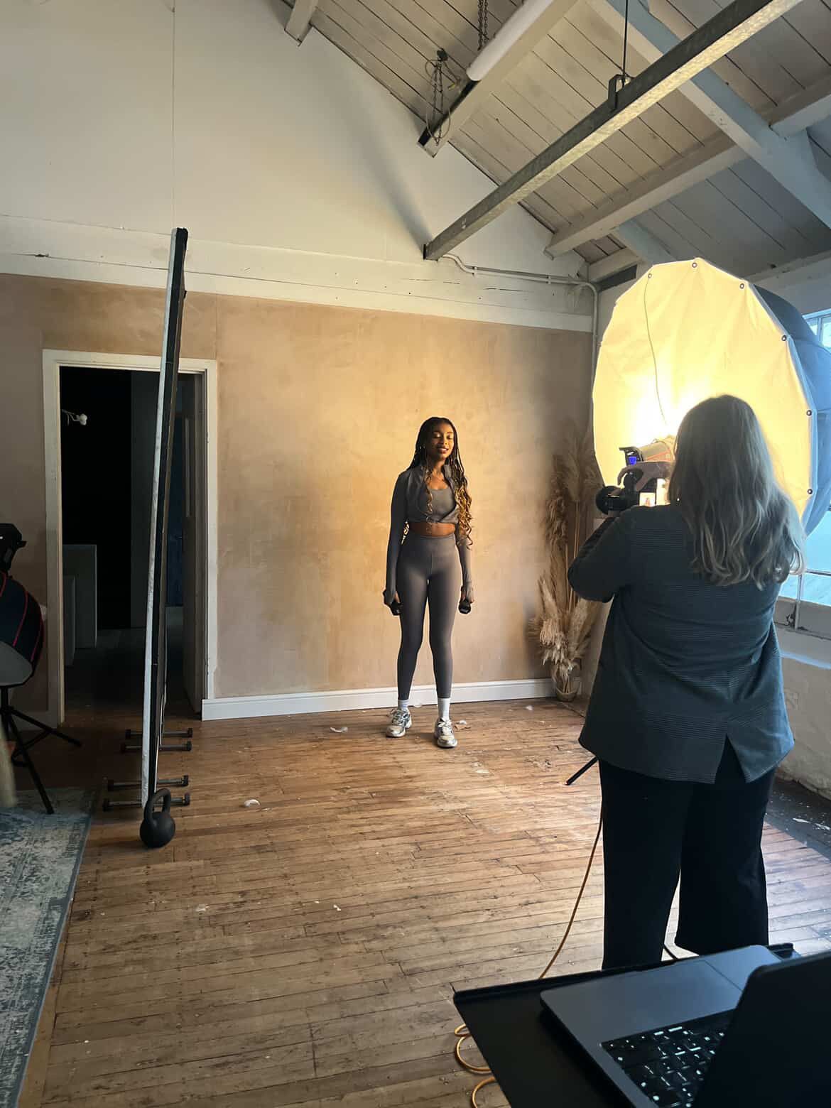 Photographer using large reflector to photograph woman in gray outfit in studio.
