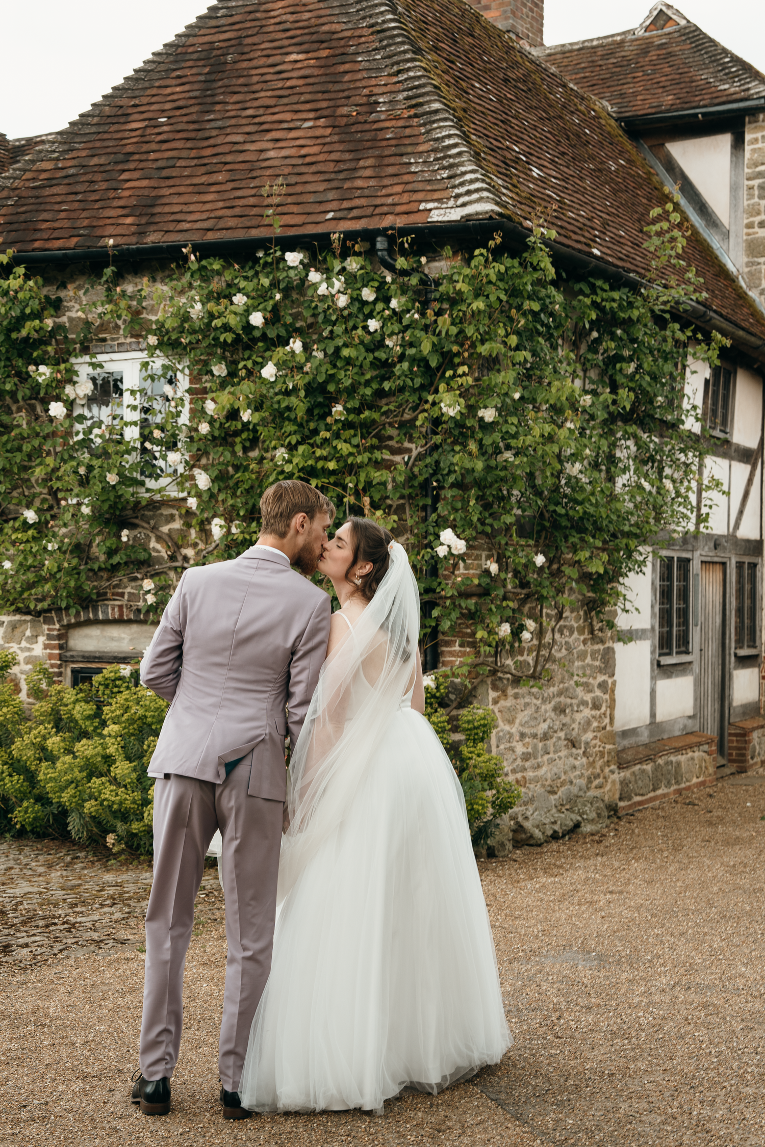 A bride and groom share a romantic kiss outside a charming stone and timber English country house adorned with white roses.