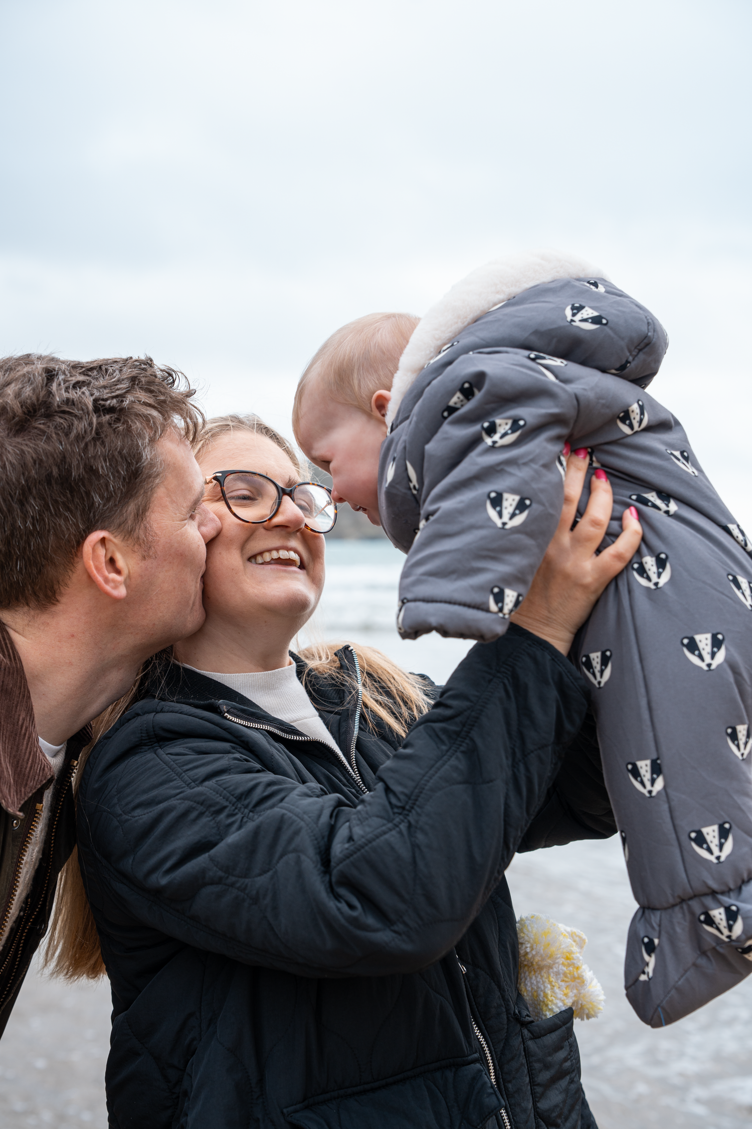 Parents kissing and lifting laughing baby in grey patterned snowsuit on a cloudy beach day.
