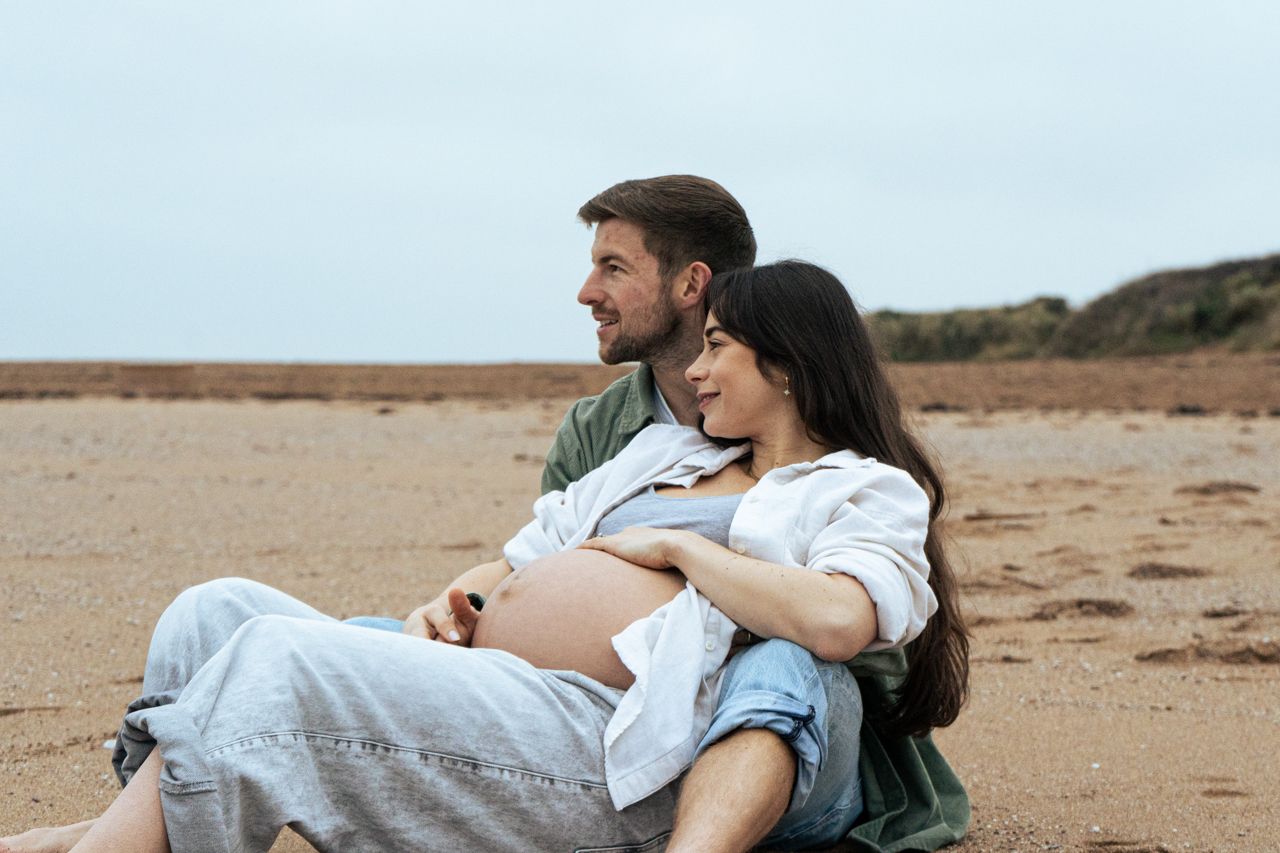 Couple sitting together on sandy beach, pregnant woman leaning against partner while gazing at ocean.