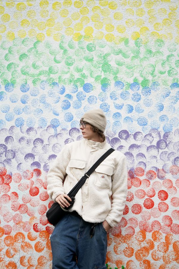 Person in beige beanie and white fleece jacket standing before vibrant rainbow polka dot wall.