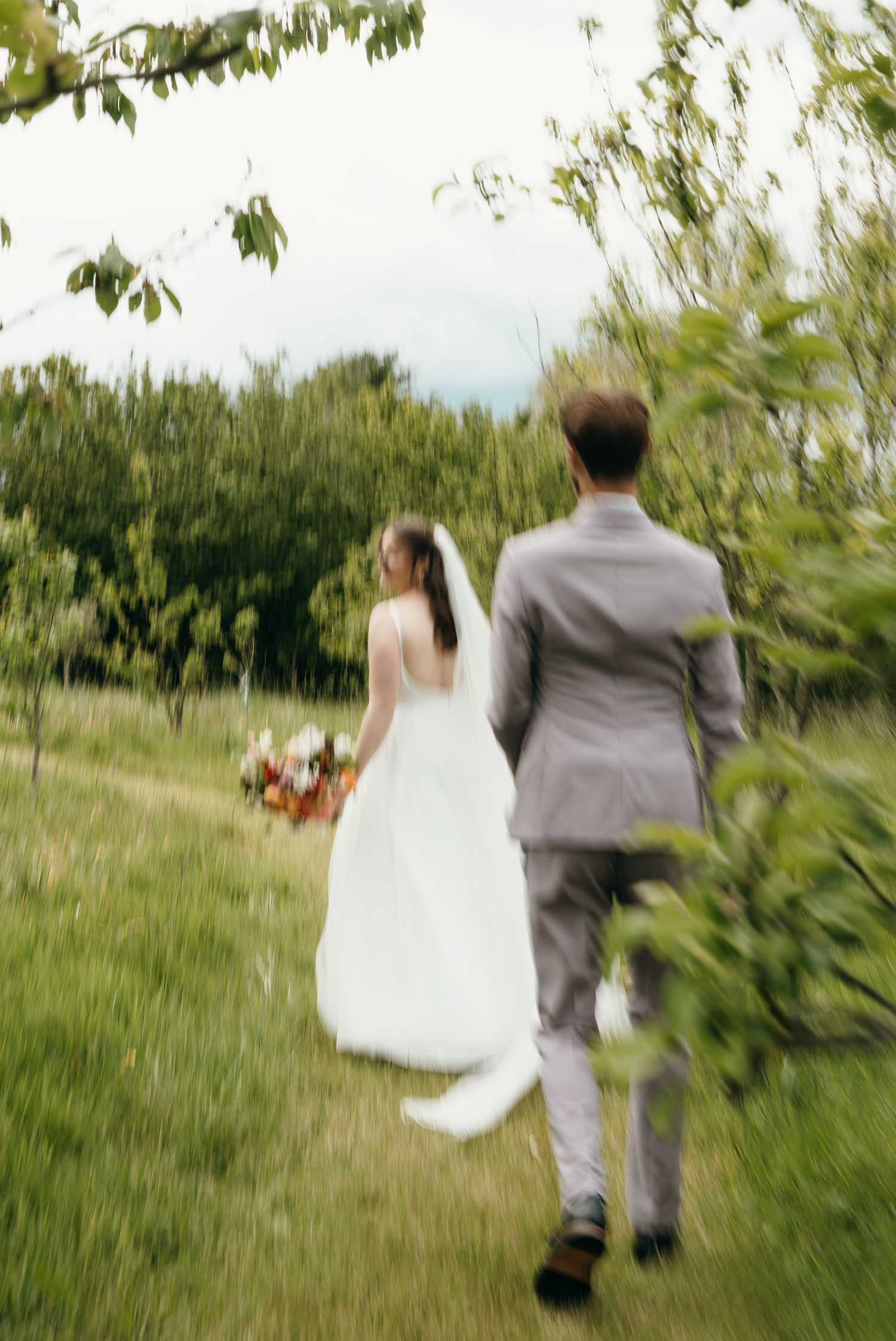 A bride in white gown and groom in gray suit walk hand-in-hand through a lush green garden on their wedding day.