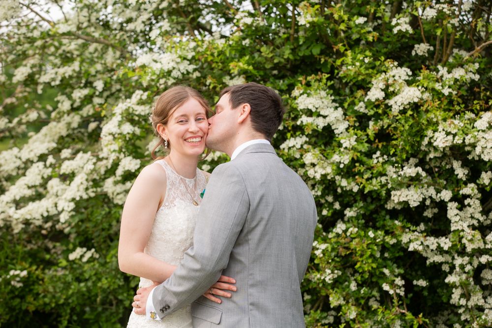 Groom kissing brides cheek as she smiles in front of blooming white flowering trees in garden.