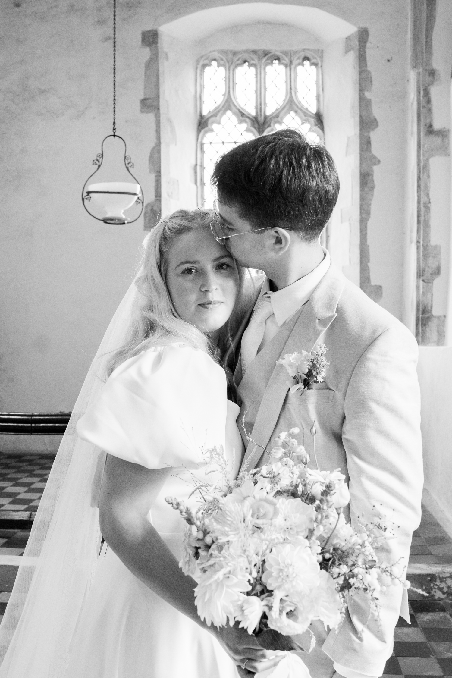 Black and white portrait of bride and groom embracing in church holding white bouquet.
